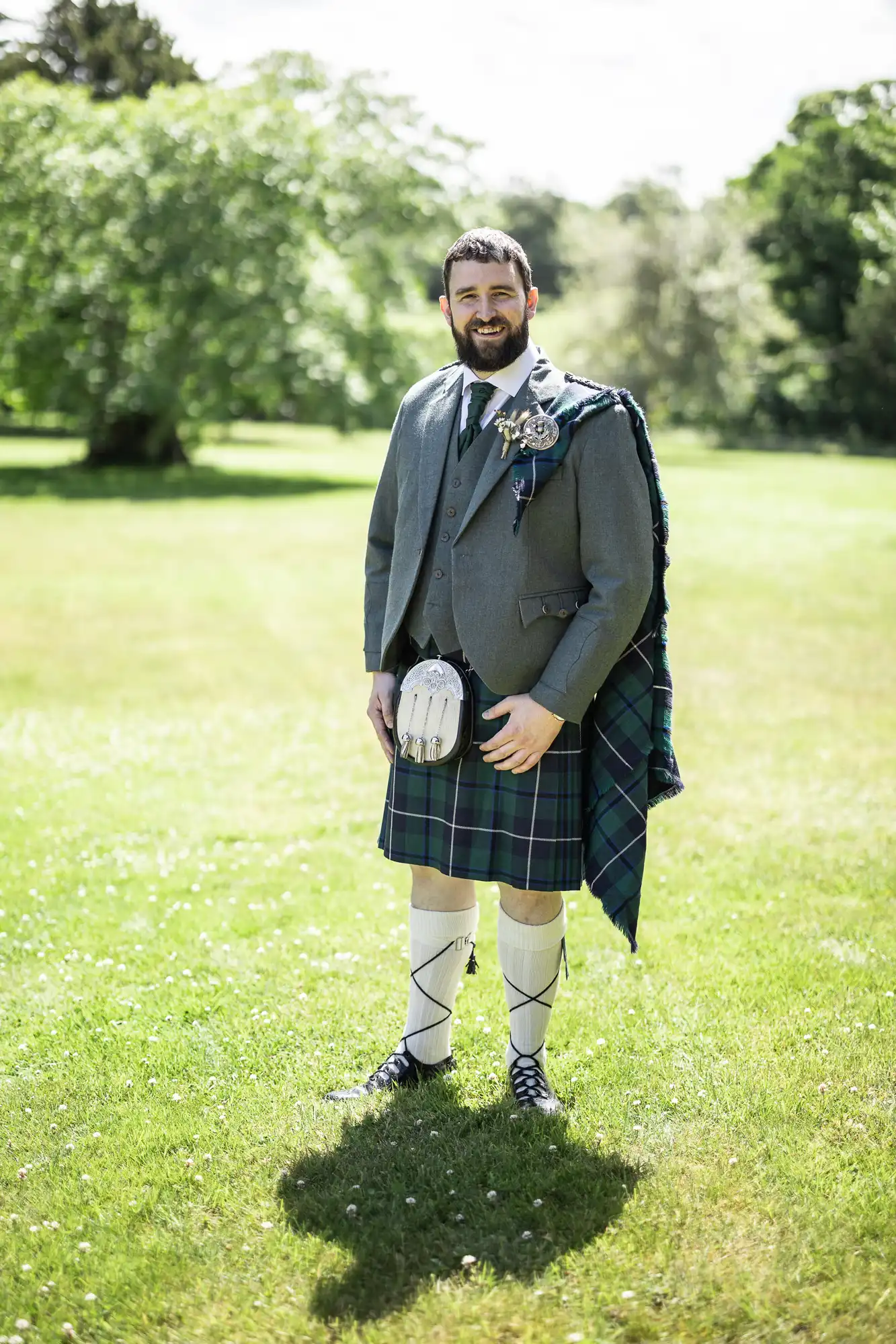 A man with a beard stands on green grass outdoors, smiling at the camera. He wears a traditional Scottish outfit with a grey jacket, green tartan kilt, sporran, knee-high socks, and black shoes. Trees are visible in the sunlit background.