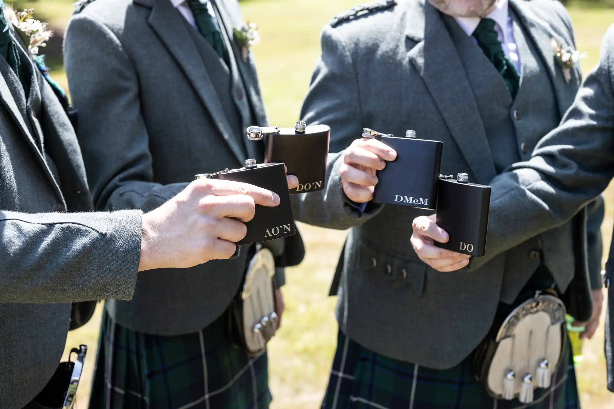 Four people dressed in grey jackets, waistcoats, and green tartan kilts hold up black flasks with white initials. Their faces are only partially visible. Each person wears a sporran and buttonhole, suggesting a formal or celebratory Scottish event.
