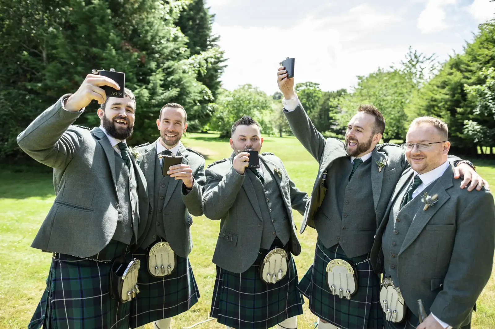 Five men stand outside on grass, smiling and wearing matching grey jackets, ties, and blue-green tartan kilts. Three hold up mobiles, possibly for selfies, while another drinks from a hip flask. They appear cheerful and celebratory, surrounded by trees and sunlight.