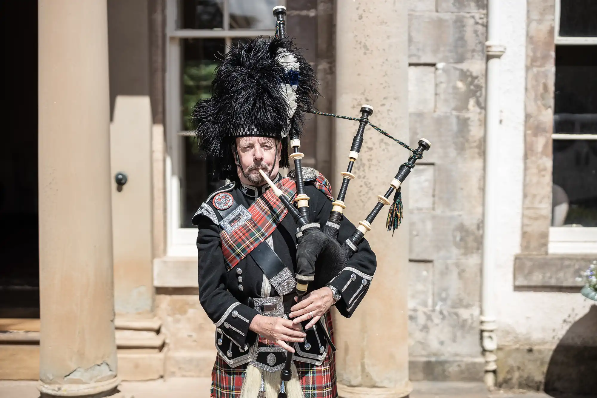 A man in traditional Scottish attire, including a tartan kilt, plaid sash, and feathered hat, stands outdoors in front of stone columns and a window, playing the bagpipes. Sunlight highlights his uniform's details and the instrument’s pipes.