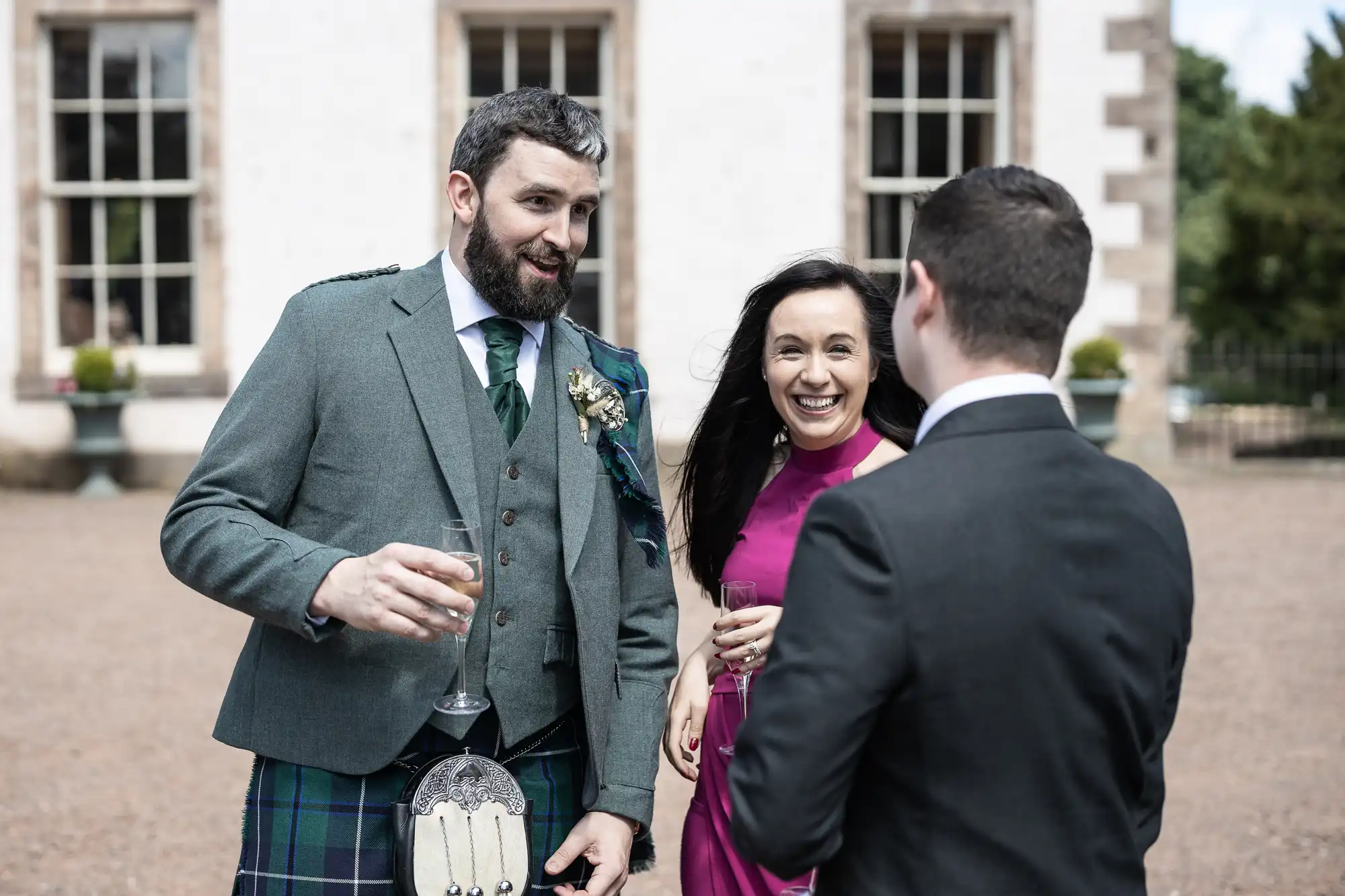 Three people are chatting and smiling outdoors at a formal event. One man wears a kilt and grey jacket, another man wears a dark suit, and a woman in a sleeveless magenta dress holds a glass. They stand in front of a white building with large windows.