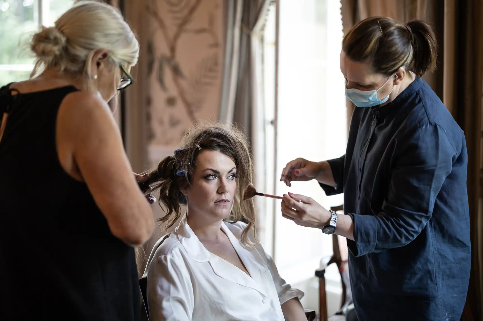 A woman in a white shirt sits while two stylists work on her; one applies make-up with a brush, wearing a face mask, and the other styles her hair with rollers. The setting appears to be indoors with soft natural light and neutral curtains.