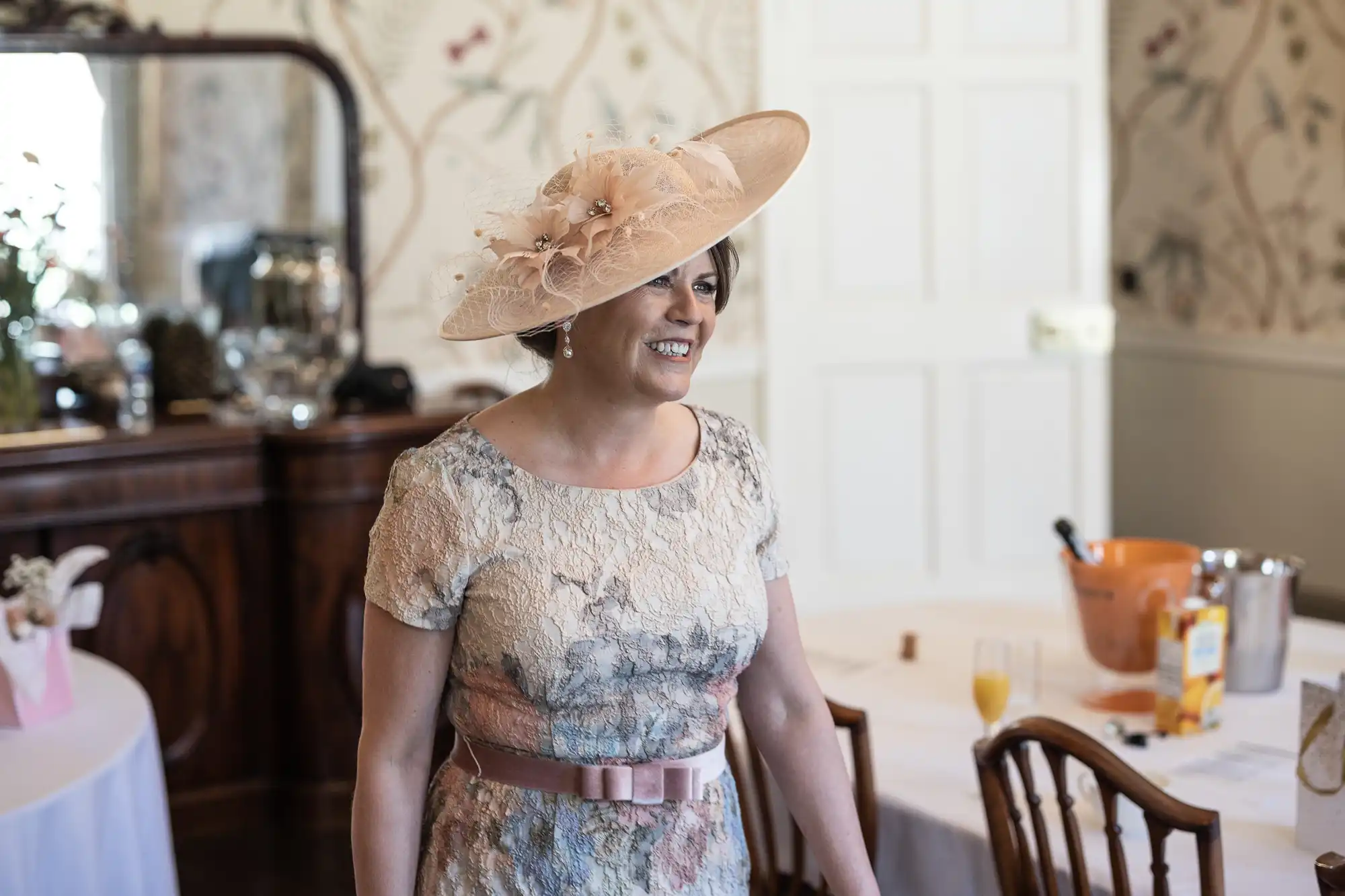 A woman in a pale, floral dress and a wide-brimmed hat decorated with feathers smiles indoors. She stands near a table with glasses and a bucket of drinks. The room has floral wallpaper, wooden furniture, and soft natural lighting.