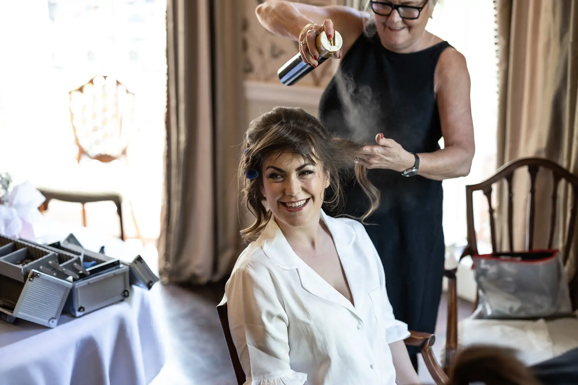 A smiling woman with curled hair sits in a white shirt while another woman sprays her hair with hairspray in a well-lit room. There are open hair styling tools on a nearby table and elegant curtains and chairs in the background.