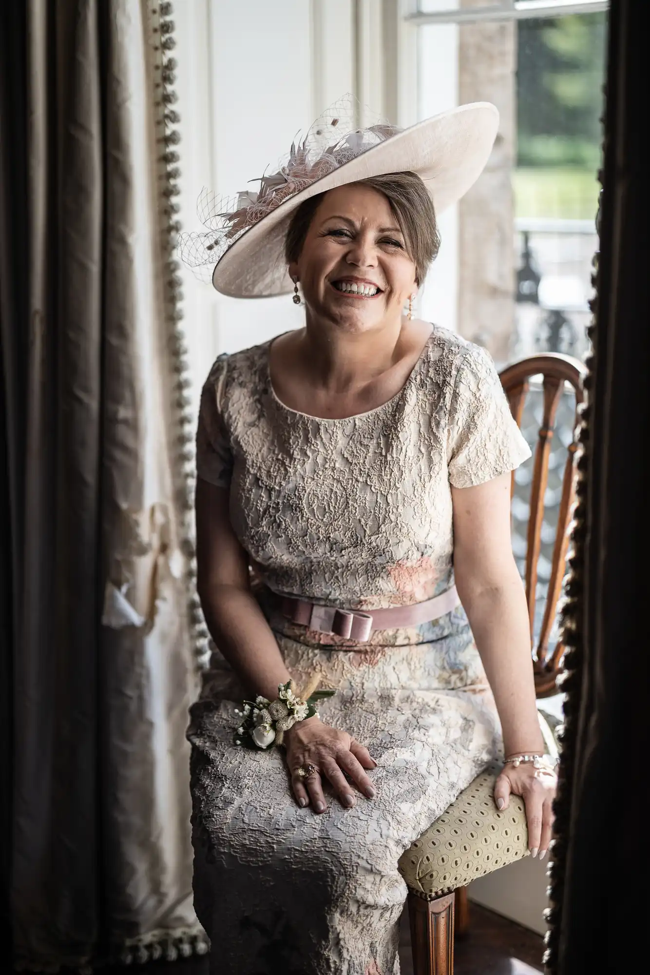 A smiling woman in a lace dress and wide-brimmed hat sits on a cushioned chair by a window with curtains. She wears a corsage, belt, and jewellery, with sunlight illuminating her face. The background shows a glimpse of greenery and an iron railing outside.