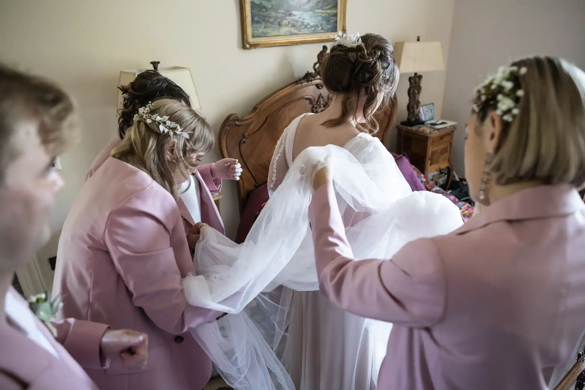 A bride in a white gown stands in a bedroom as four bridesmaids in pink suits, adorned with floral headpieces, carefully arrange her long veil. The setting is intimate, with soft lighting and a wooden bed and lamp in the background.