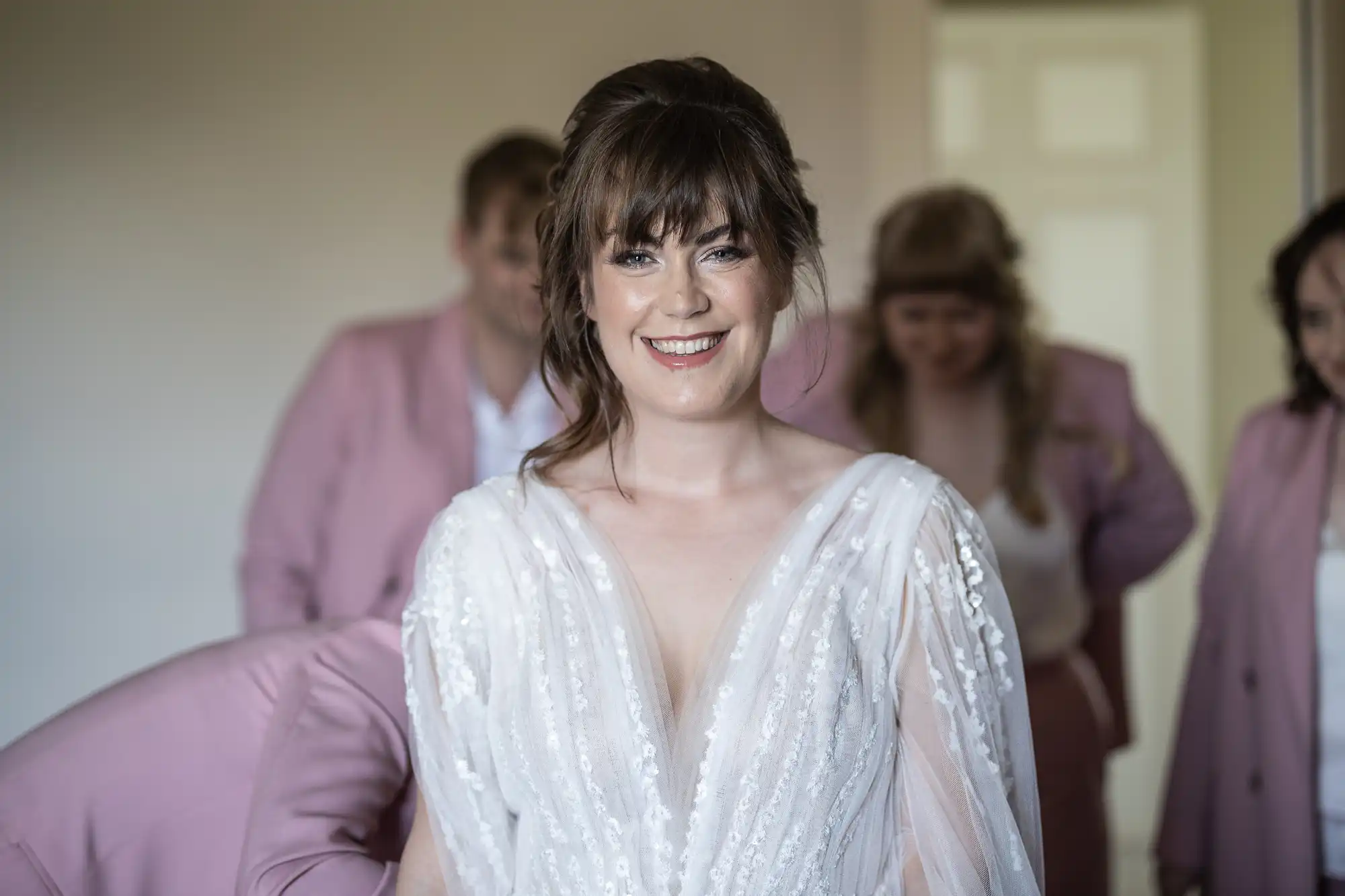 A bride with brown hair and a fringe smiles, wearing a white, textured, V-neck wedding dress. Behind her, three bridesmaids in pink outfits are slightly out of focus, standing in a softly lit room with a white door in the background.