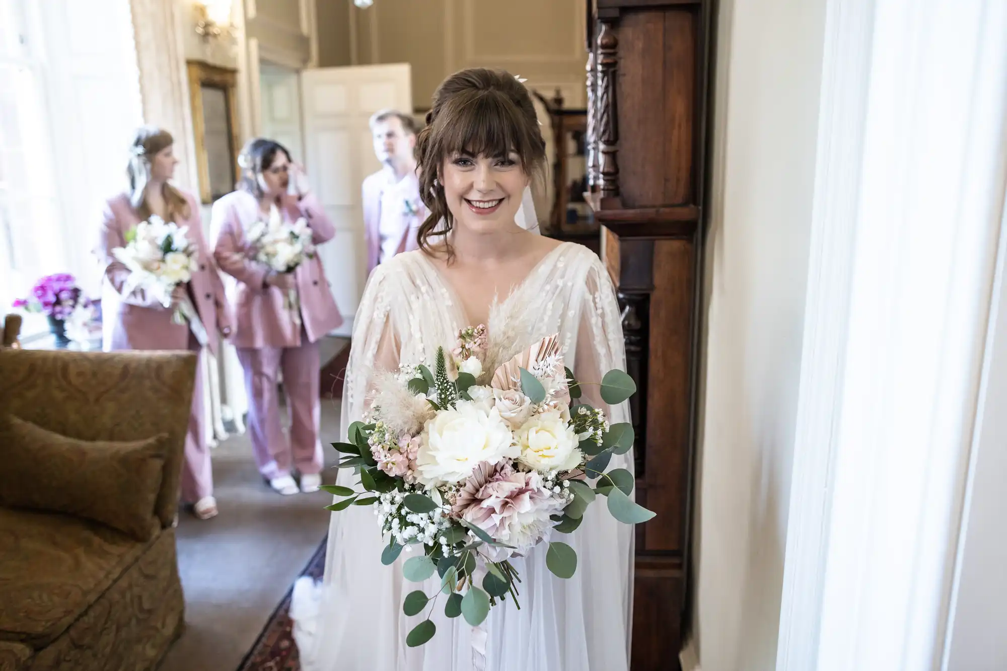 A bride in a white dress with a large bouquet of white and pink flowers smiles at the camera. Behind her, three bridesmaids in matching light pink suits and another woman in a mauve dress hold bouquets, standing in a bright, elegant room.