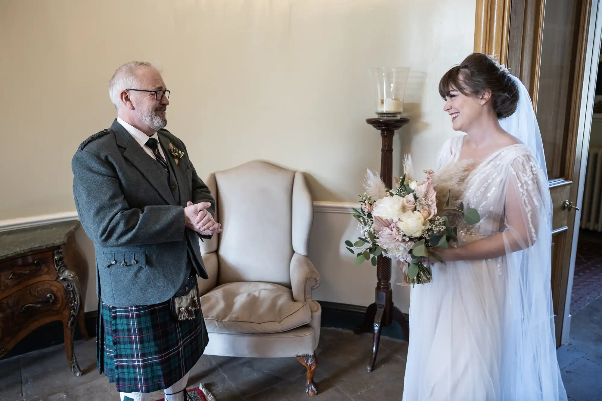 A smiling older man in a grey jacket and traditional Scottish kilt admires a bride in a white gown holding a bouquet. They stand indoors near a cream armchair and a wooden table, with soft natural light illuminating the scene.