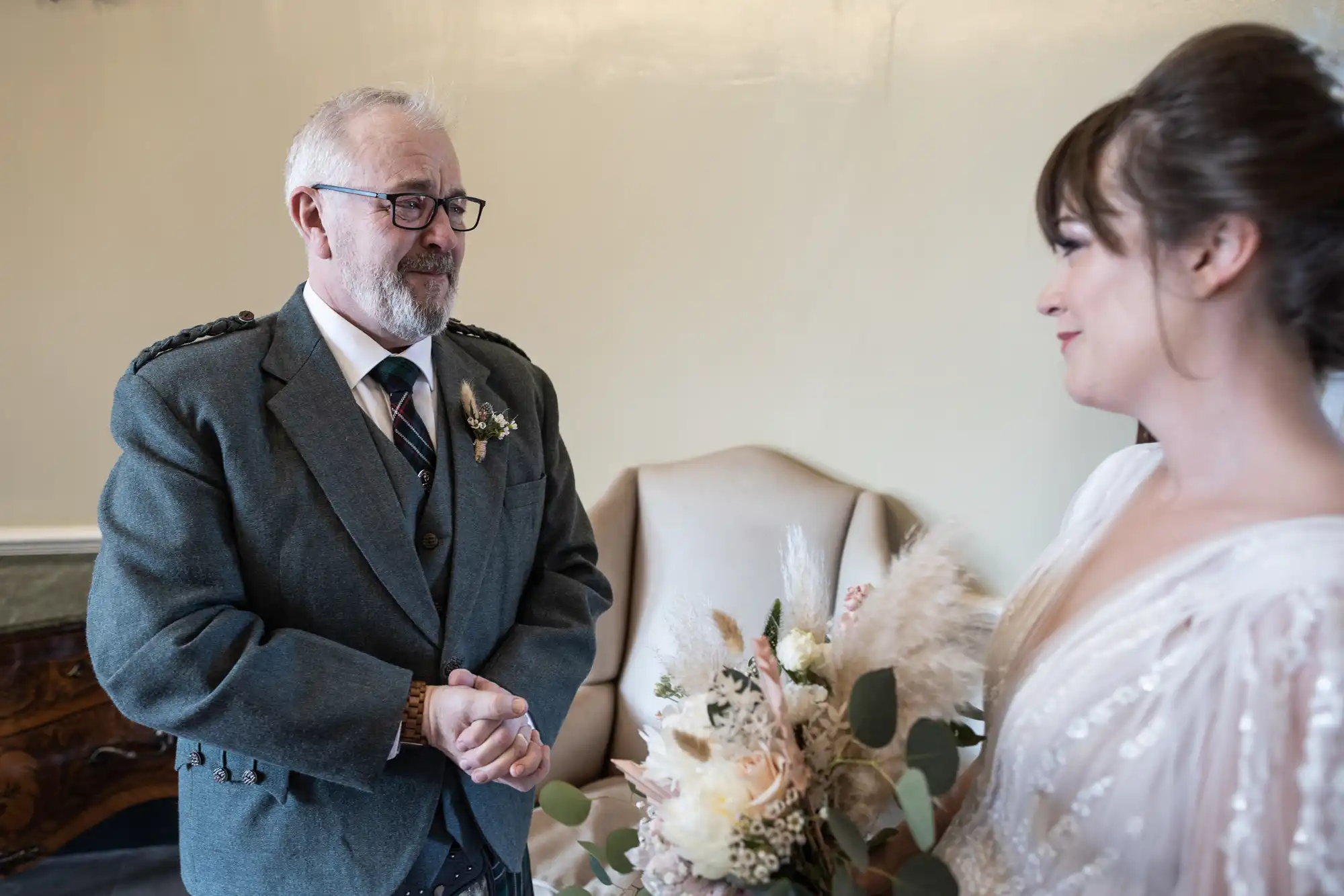 An older man in a grey suit with a buttonhole smiles warmly at a bride in a white dress holding a bunch of flowers. They stand indoors near a light-coloured armchair, sharing a tender moment before the wedding ceremony.