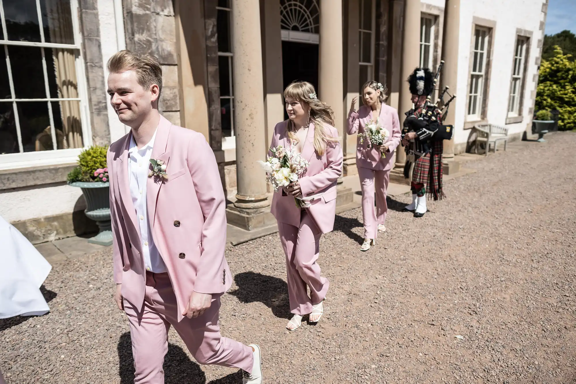 Three people in matching light pink suits, holding bouquets, walk in a line outside a grand stone building. A bagpiper in traditional Scottish attire follows them. The sky is clear and the scene is bright and cheerful.