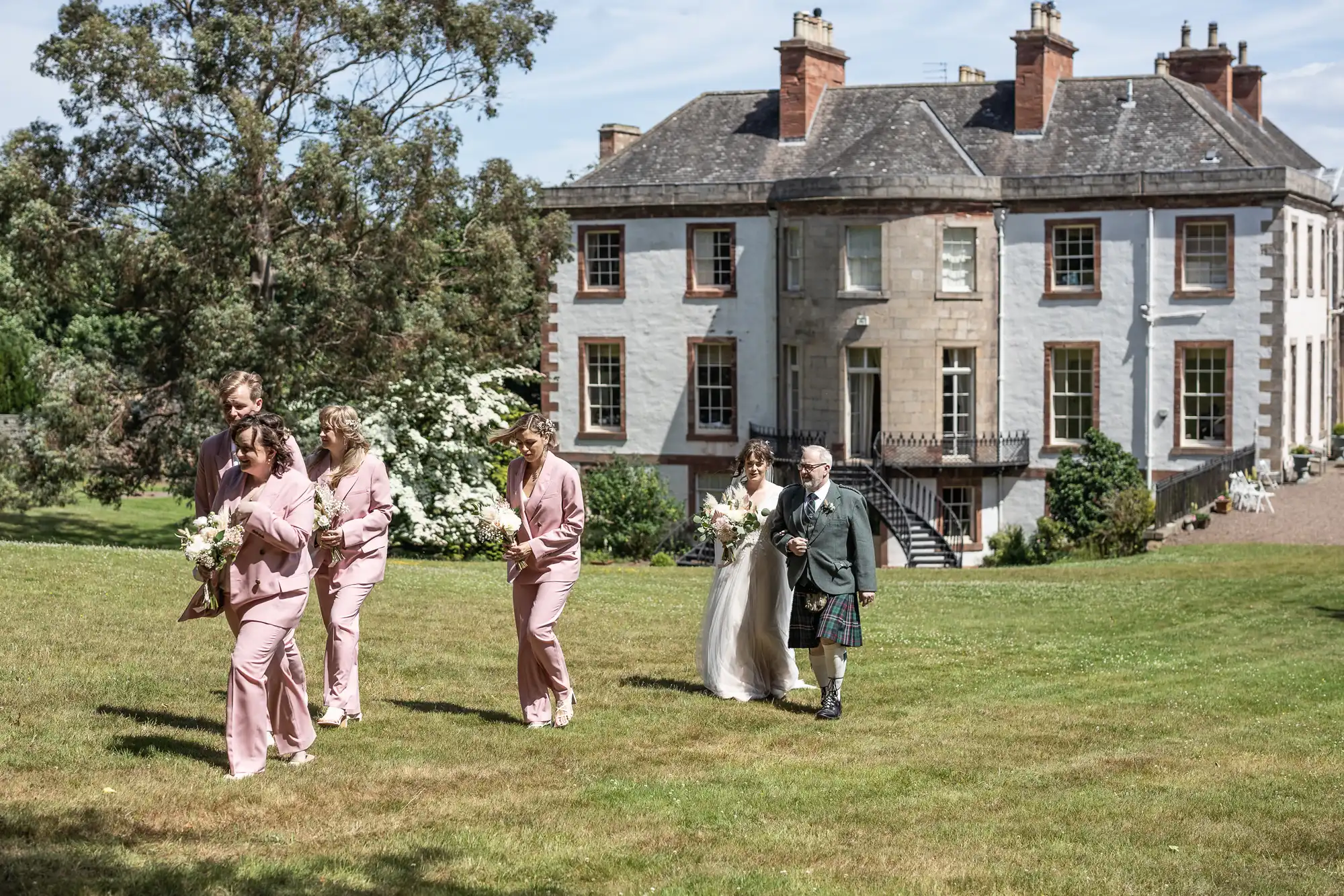 A bride in a white dress walks on a sunny lawn with an older man in a kilt beside her. Ahead, five bridesmaids in matching pink suits hold bouquets. Behind them is a large, elegant two-storey house surrounded by greenery under a blue sky.