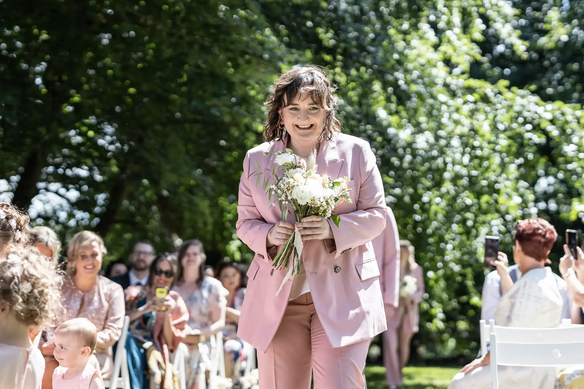 A person in a light pink suit holds a bouquet of white flowers, walking down an outdoor aisle lined with seated guests. Sunlight filters through lush green trees, casting dappled shadows on the scene. Some guests are smiling and taking photos.