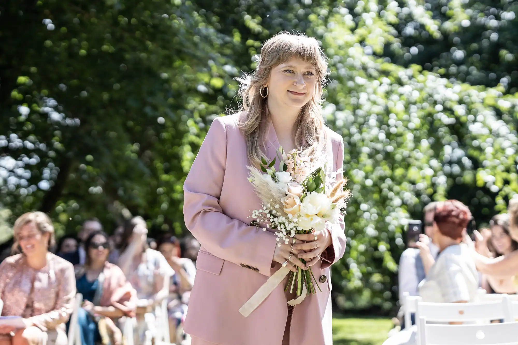 A person with light brown hair wearing a pale pink suit jacket holds a bouquet of white and pastel flowers, walking outdoors at a sunny wedding ceremony. People seated in rows of white chairs and green trees are visible in the background.