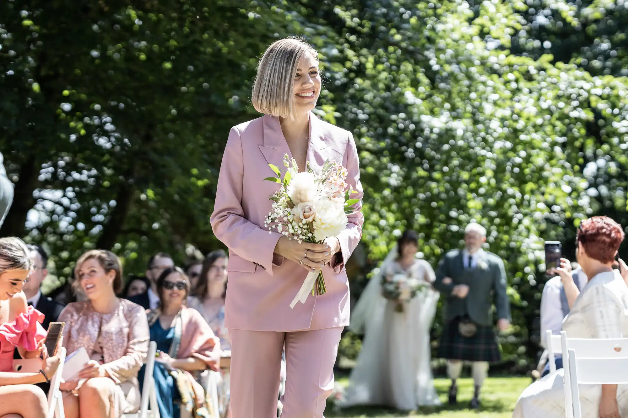 A smiling woman in a light pink suit holds a bouquet of flowers while walking outdoors at a wedding event. Seated guests watch her, and lush green trees fill the background. Some people, including a bride in white, stand blurred in the distance.