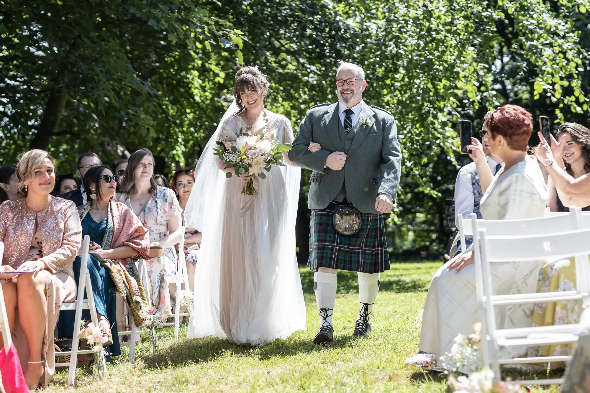 A smiling bride in a flowing white dress and veil walks down an outdoor aisle with an older man in a kilt. Guests, seated on white chairs, watch and take photos. Sunlight filters through leafy trees, creating a bright, cheerful atmosphere.