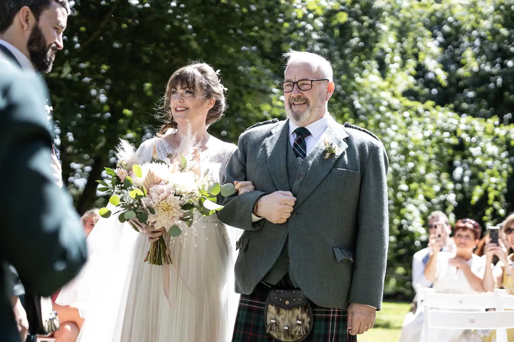 A bride in a white dress and holding a bouquet walks arm-in-arm with an older man in a kilt and jacket. They smile as they walk outdoors in the sun, surrounded by greenery and guests seated on white chairs, some taking photos.