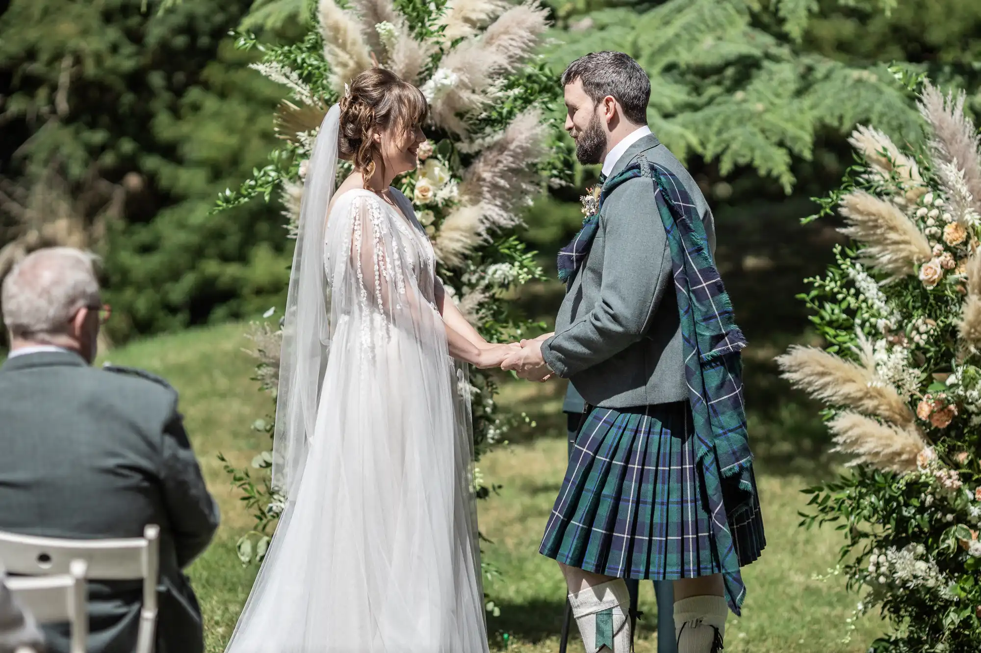 A bride in a white dress and veil holds hands with a groom in a traditional Scottish kilt during an outdoor wedding ceremony. They stand facing each other, framed by pampas grass and greenery, with a guest seated nearby.