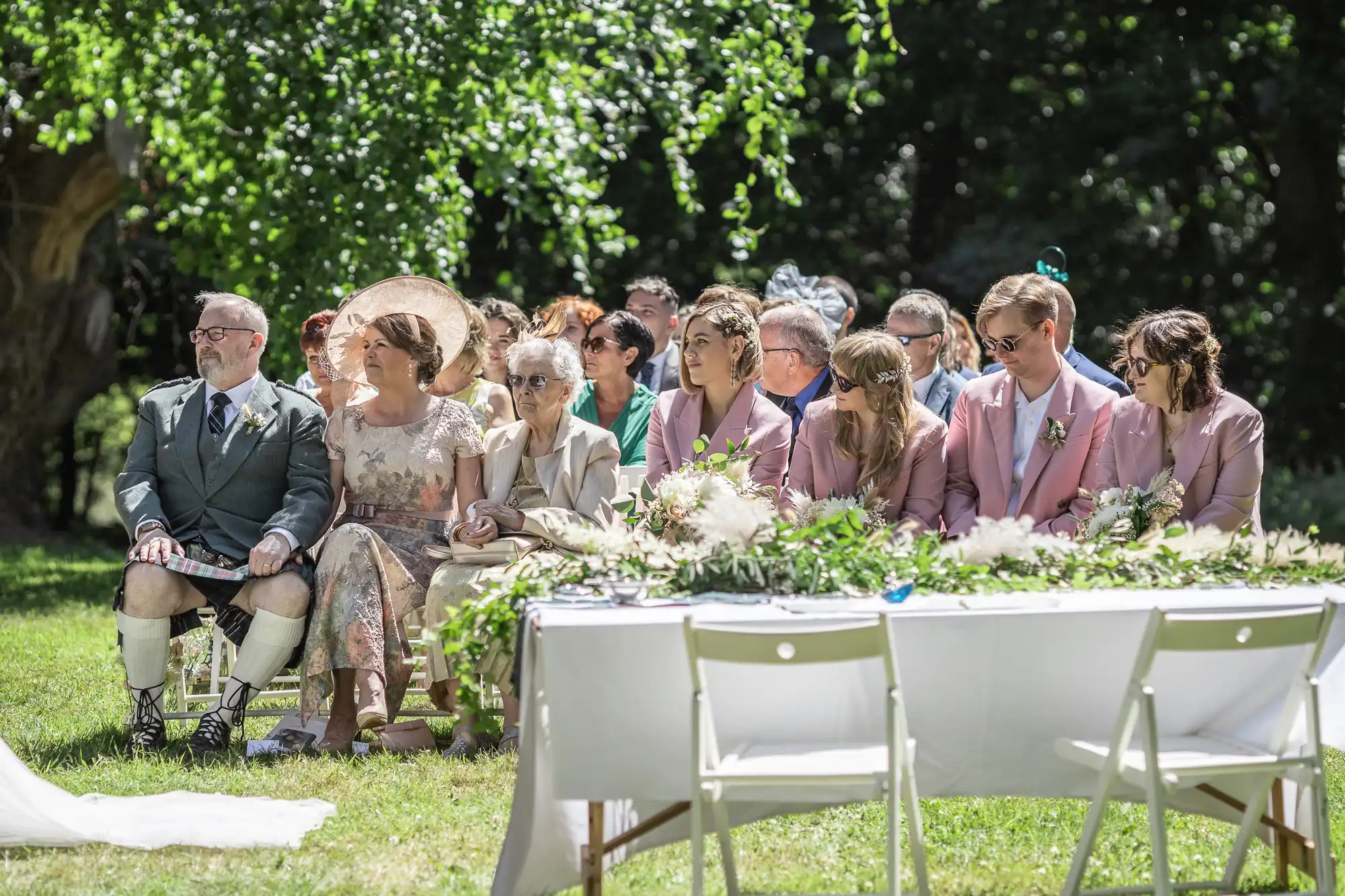 A group of wedding guests, dressed in formal attire, sit on chairs outdoors under a tree. Some wear pastel colours and floral patterns. A table with greenery and white flowers is in the foreground. Sunlight filters through the leafy background.