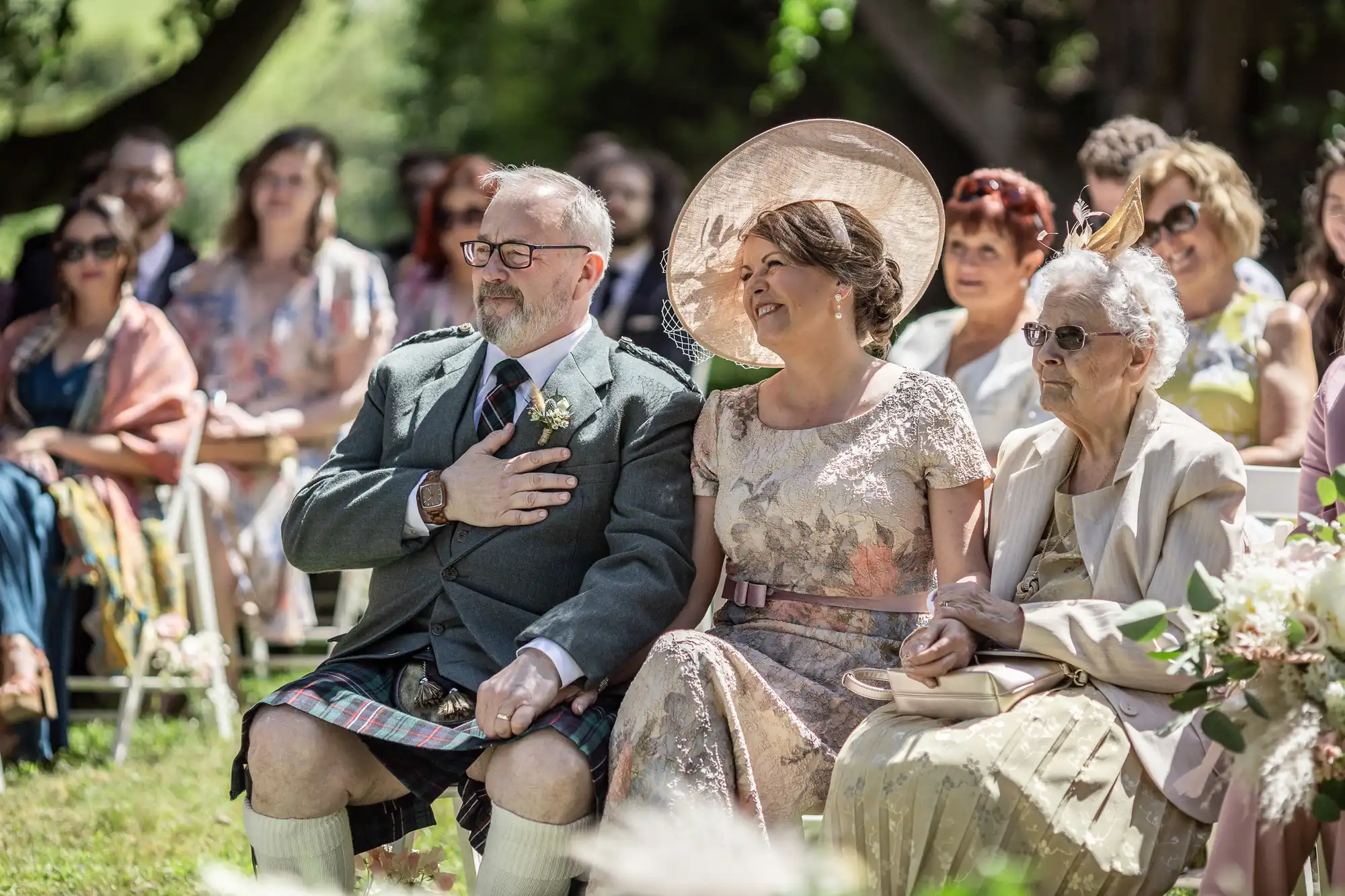 A group of wedding guests sits outdoors, smiling and watching the ceremony. In front, an older woman in white, a man in a kilt with his hand on his chest, and a woman in a hat holding his hand. Other guests are seated behind them amid greenery and sunlight.