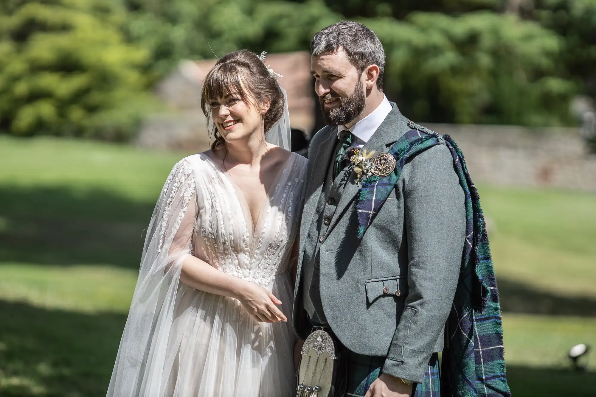 A bride in a white, flowing gown with beaded details stands beside a groom in a grey jacket, tartan sash, and kilt. Both smile and stand outdoors on green grass, with blurred trees and stone wall in the background, sunlight illuminating them.