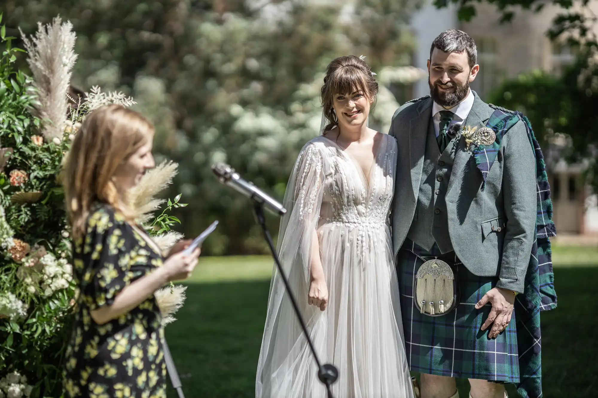 A bride in a white dress and a groom in a kilt stand smiling outdoors during their wedding ceremony, listening to a woman speaking at a microphone. Lush greenery and floral arrangements decorate the background.