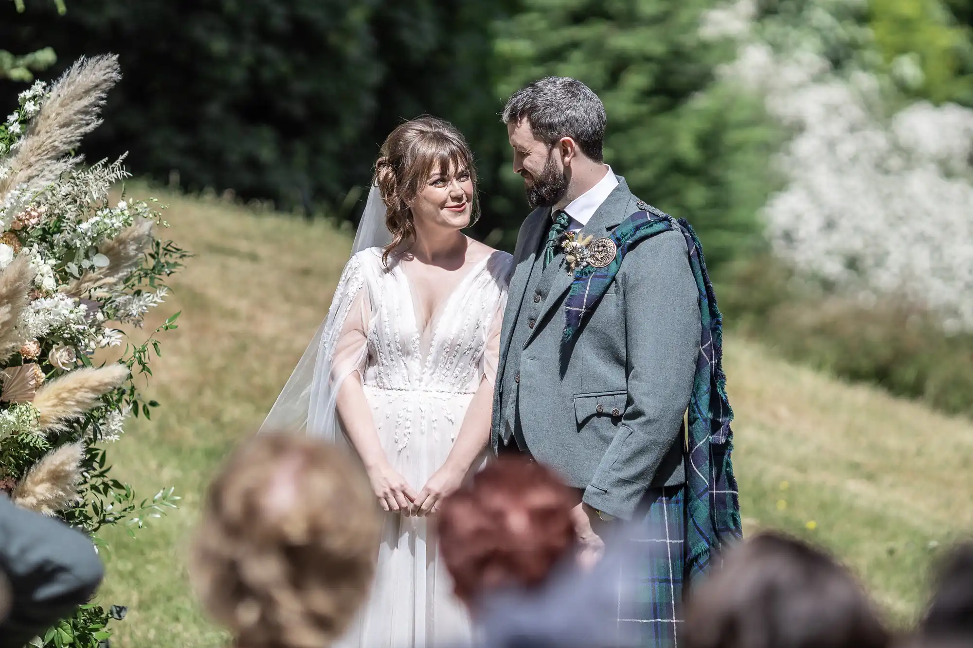 A bride in a white dress and veil stands smiling beside a groom in a grey suit with a tartan sash. They hold hands outdoors near floral arrangements, surrounded by greenery, with several guests’ heads visible in the foreground.