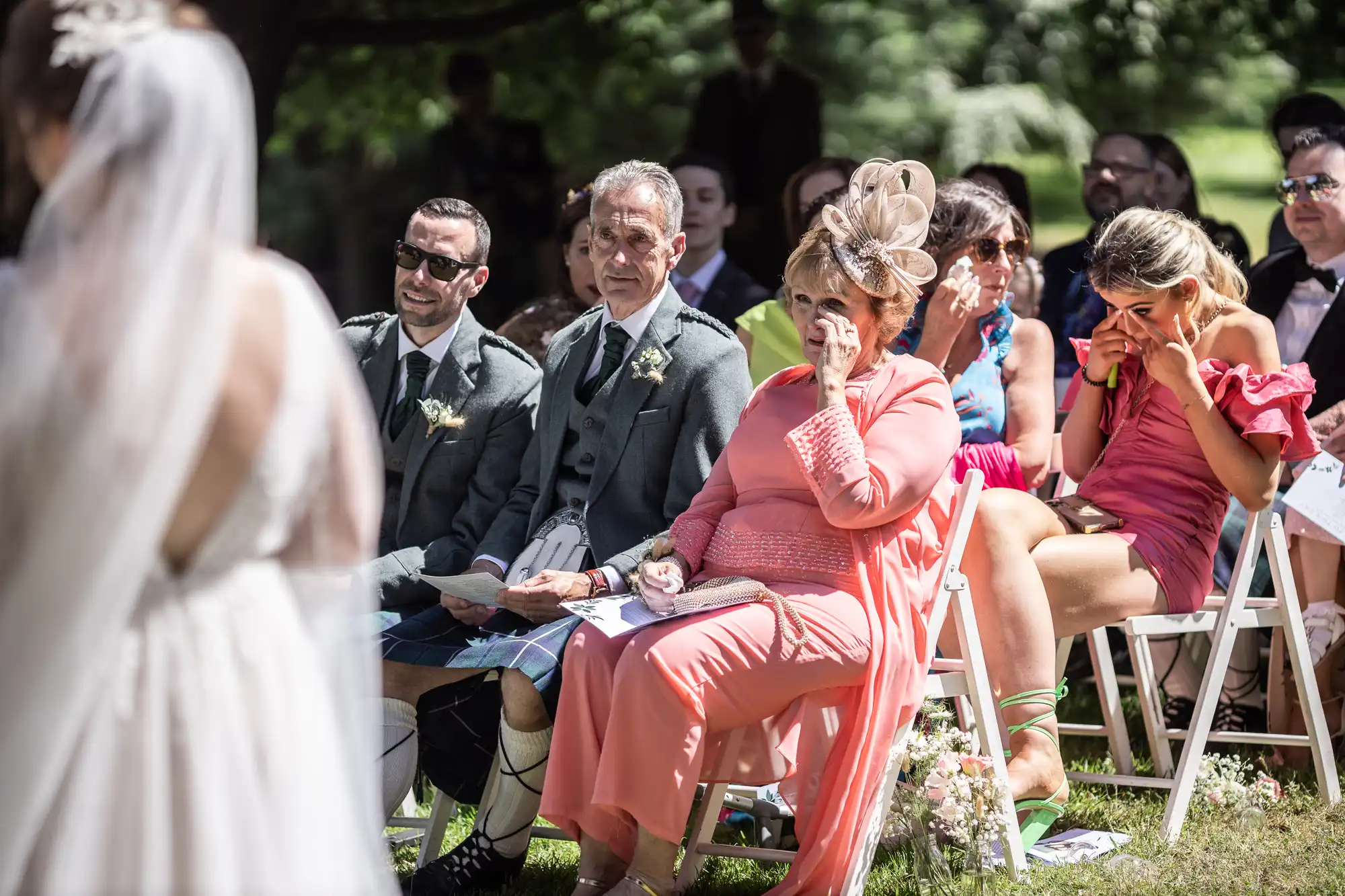 A group of wedding guests sits outdoors on white chairs, watching a bride in a white veil. Two women in front, one in coral and one in pink, wipe away tears. Men in kilts and suits are beside them, and sunlight filters through the trees onto the scene.