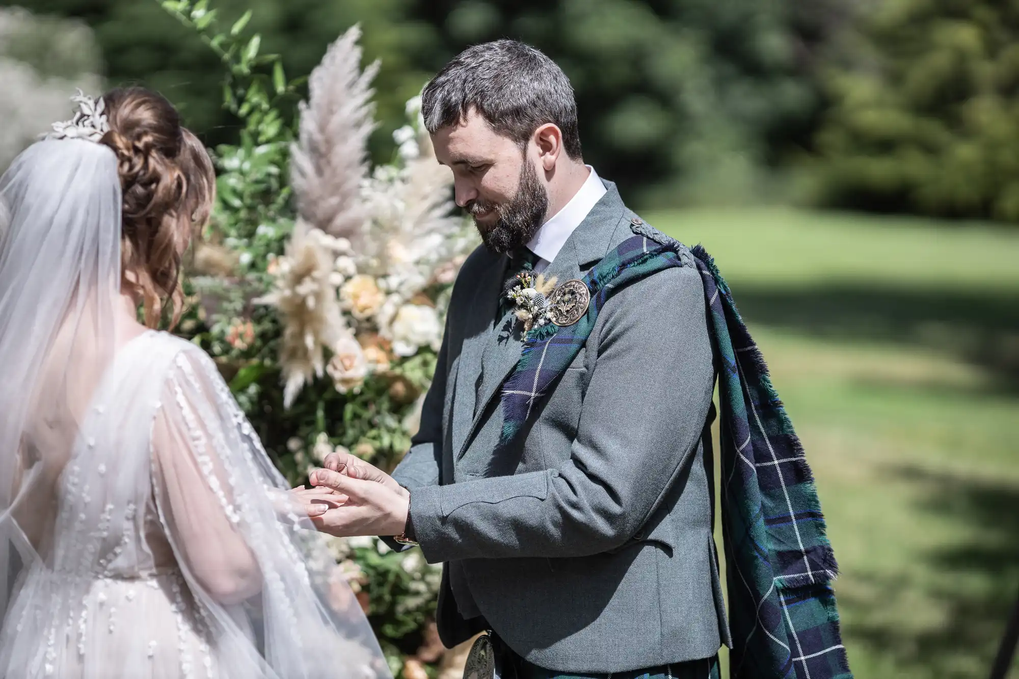 A groom in a grey suit with a tartan sash places a ring on the bride’s finger during an outdoor wedding ceremony. The bride wears a white gown with a sheer veil. Lush greenery and floral arrangements are visible in the background.