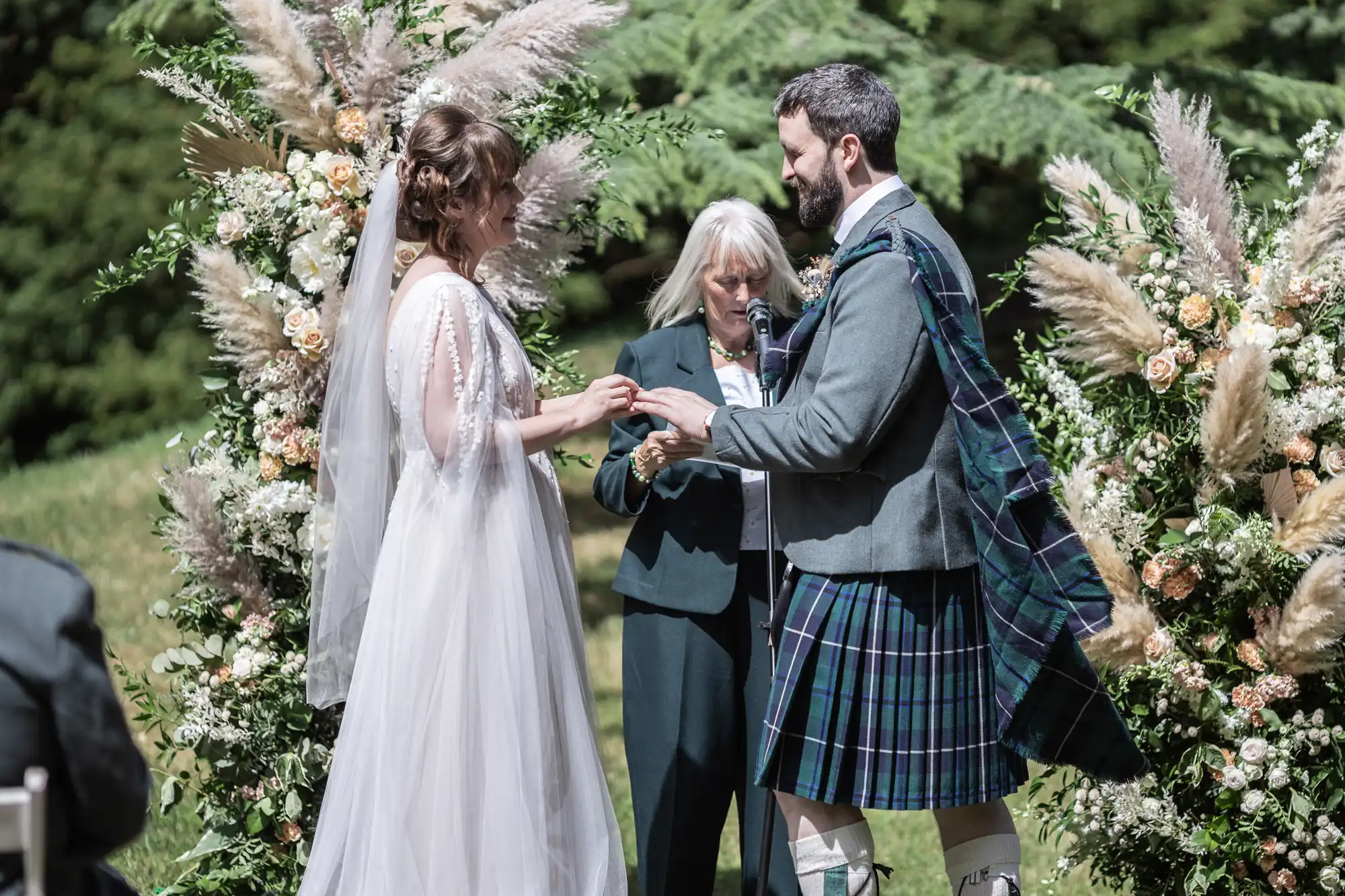 A bride in a flowing white dress exchanges rings with a groom in a traditional Scottish kilt during an outdoor wedding ceremony. They stand before a floral arch, and an officiant reads from a book. Lush greenery fills the background.