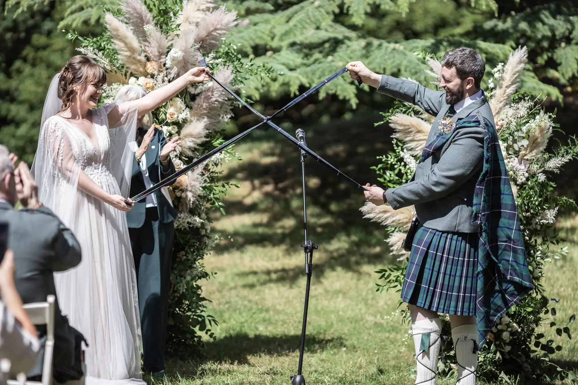 A bride in a white dress and a groom in a Scottish kilt stand outdoors facing each other, each holding a sword crossed in front of them. They smile under an arch decorated with pampas grass, surrounded by greenery and sunlight. An officiant stands behind them.