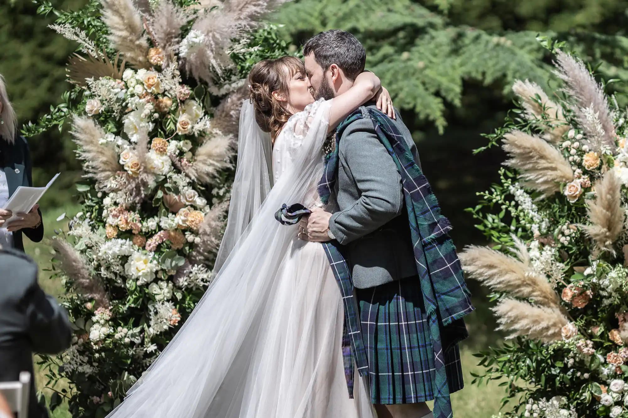 A bride in a flowing white gown and veil kisses a groom in a dark tartan kilt and jacket. They embrace outdoors, surrounded by lush greenery and two large floral arrangements with pampas grass and pastel flowers. Sunlight brightens the joyful wedding scene.