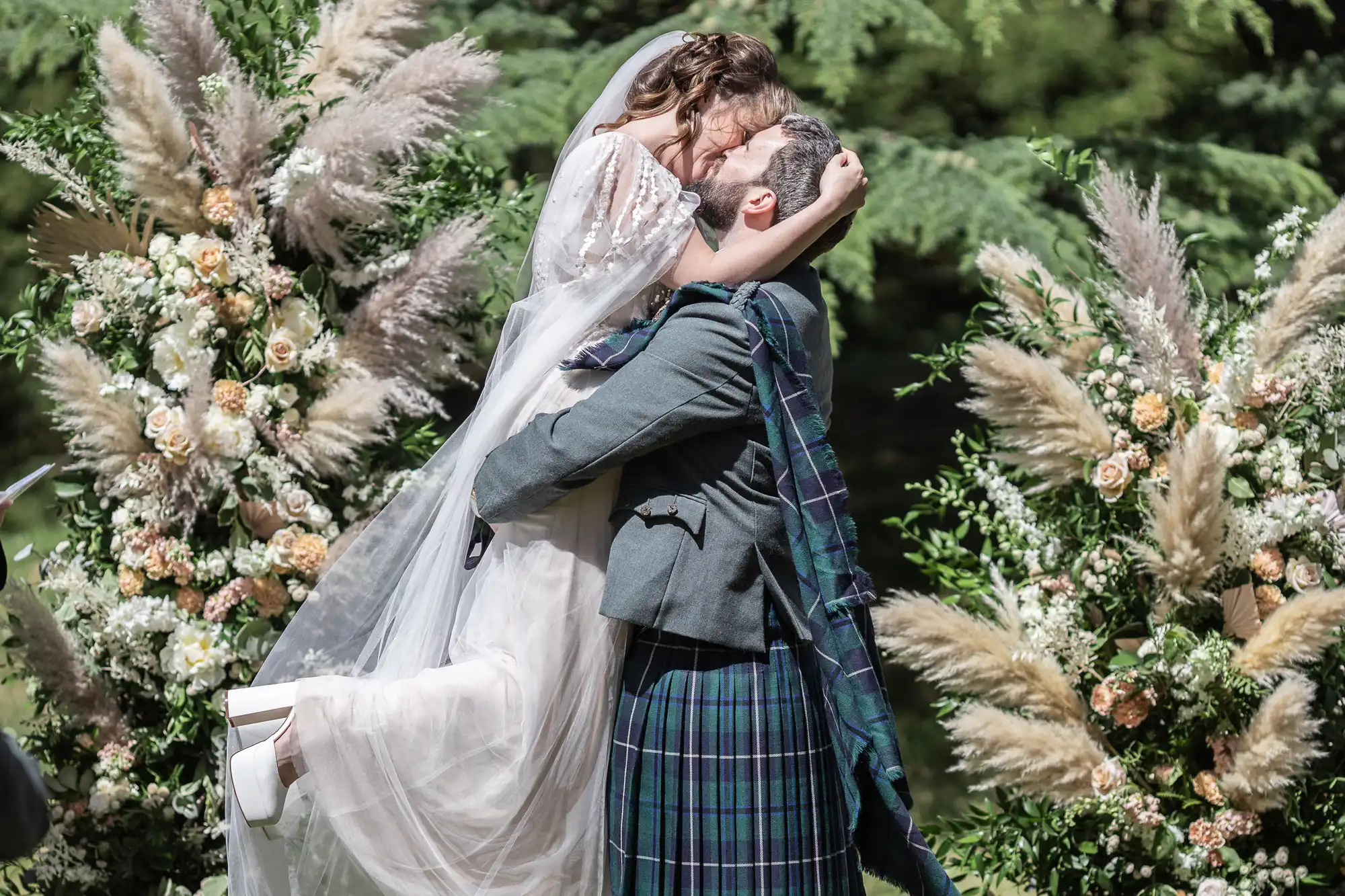 A groom in a green tartan kilt lifts a bride in a white dress and veil as they embrace outdoors. They are surrounded by lush greenery and floral arrangements with pampas grass and cream-coloured flowers, creating a joyful, whimsical wedding scene.