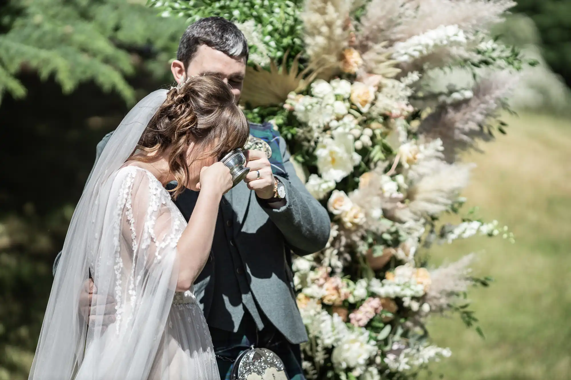 A bride and groom stand outdoors by a flower arch. The bride, in a white dress and veil, wipes her eyes with a tissue, whilst the groom, in a grey suit, gently hugs her. Sunlight falls on them and lush greenery fills the background.