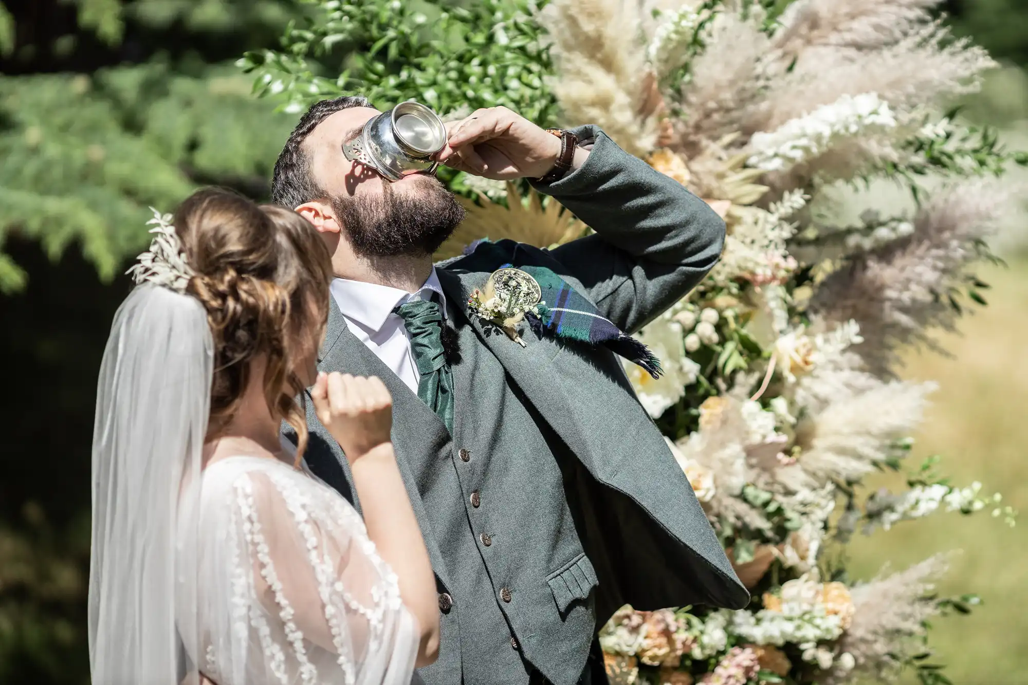 A bride in a white dress and veil stands beside a groom in a grey suit and green tie, who is drinking from a silver cup. They are outdoors, near a floral arrangement with greenery and pampas grass, in bright sunlight.