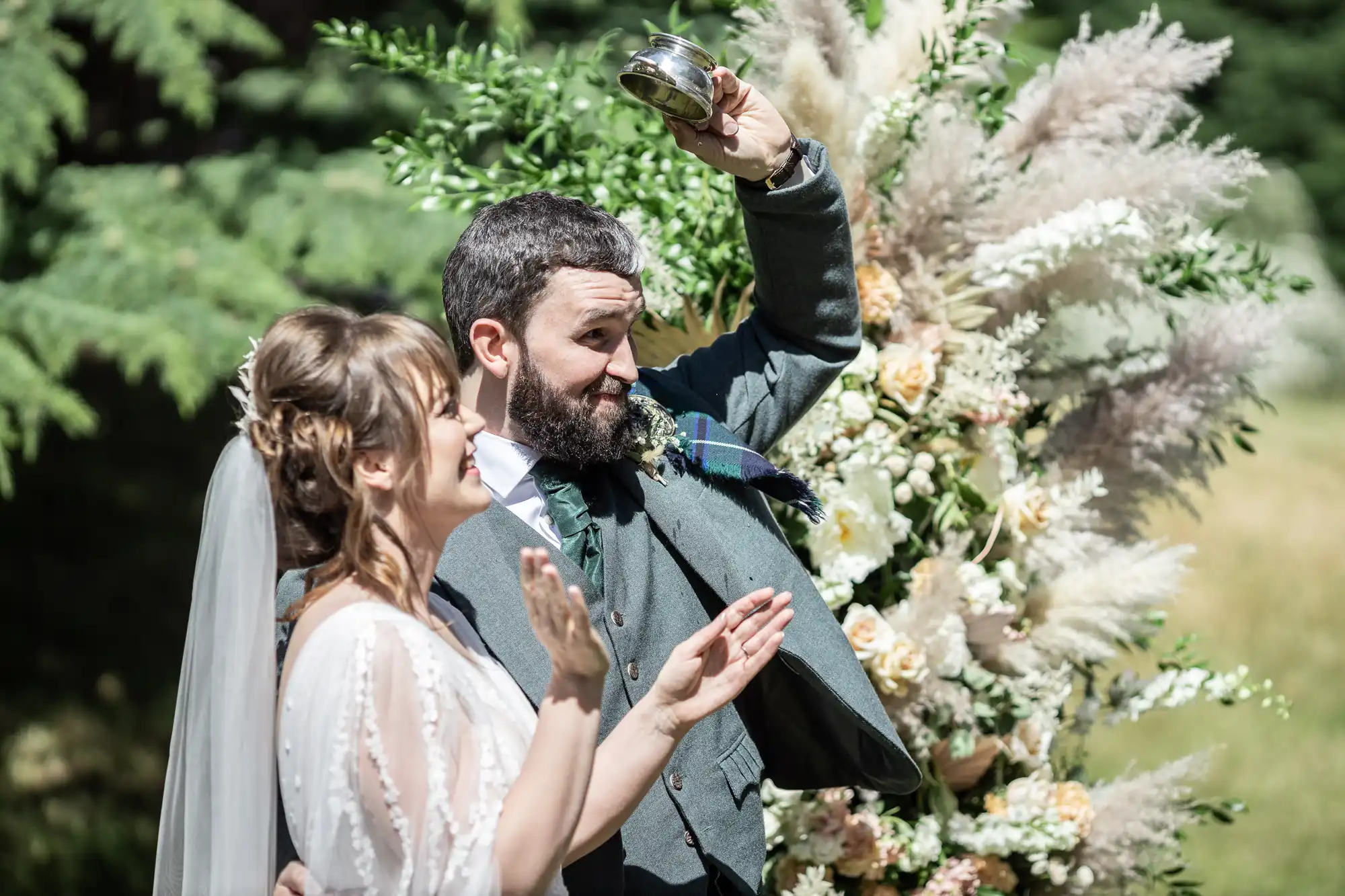 A bride and groom smile outdoors in front of lush greenery and pampas grass arrangements. The groom, in a grey suit, raises a silver cup above his head while the bride, in a white dress with a veil, claps joyfully beside him in the sunlight.