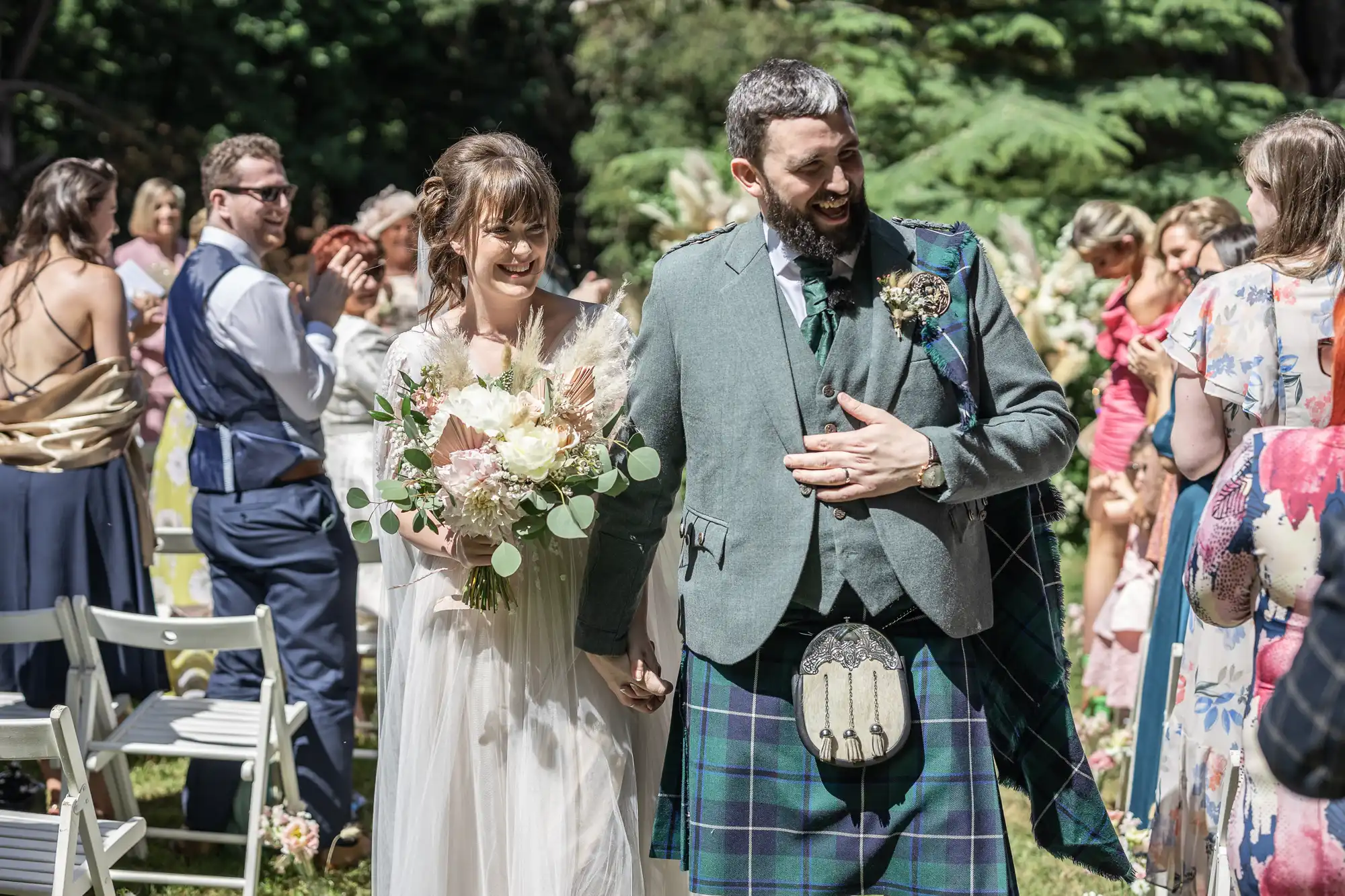 A smiling bride in a white dress holds a bouquet and walks hand-in-hand with a groom wearing a green jacket and tartan kilt. Guests, some applauding, stand and sit nearby on white chairs in a sunlit outdoor garden setting.