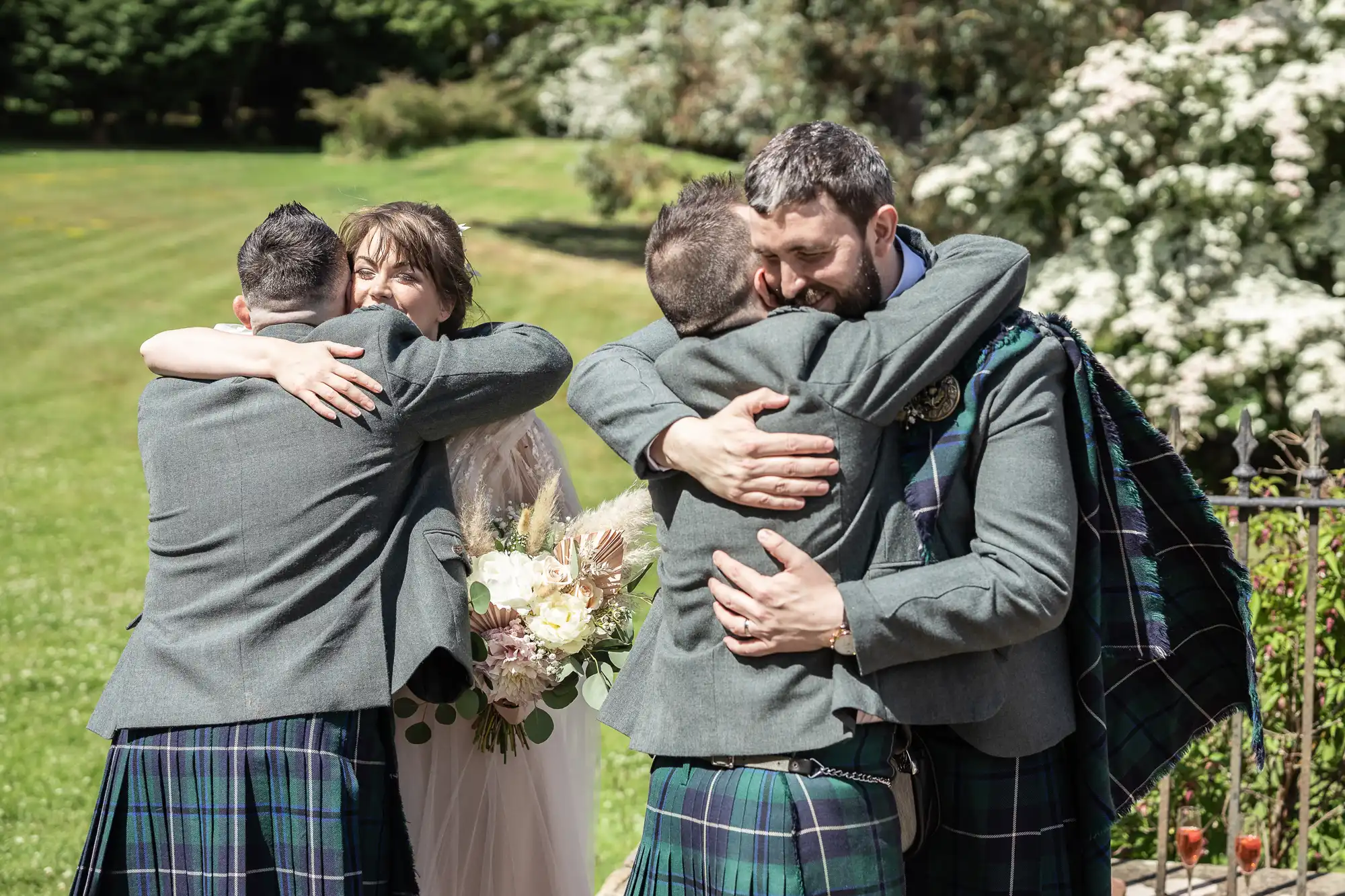 Four people wearing kilts and jackets embrace in pairs outdoors on a sunny day. A woman holding a bouquet smiles as she hugs one man; two men hug each other nearby. Lush greenery and white flowering bushes fill the background.