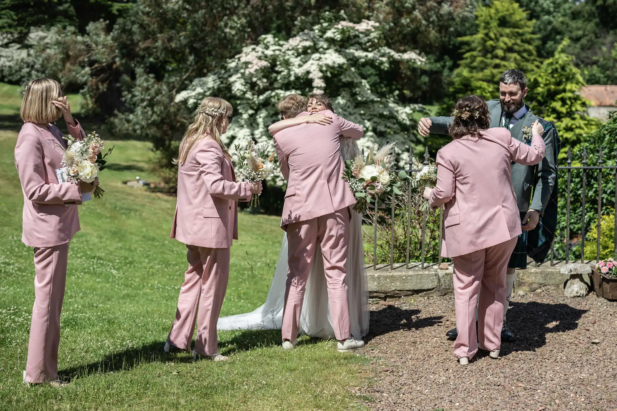 Four people in matching light pink suits, holding bouquets, gather outside on grass. One hugs a person in a white dress and veil. A man wearing a dark suit smiles nearby. Flowering bushes and green trees fill the sunny background.