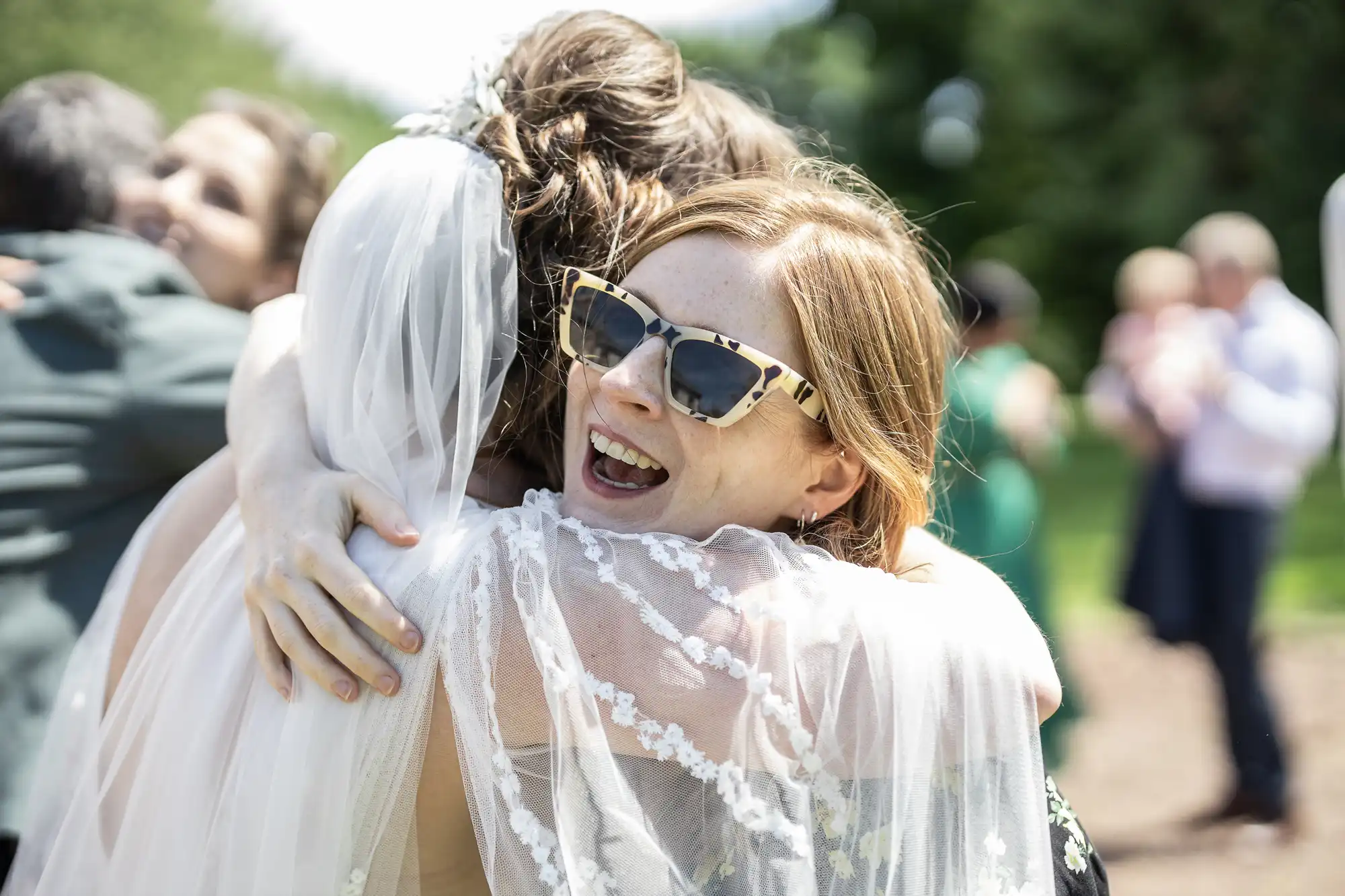 A smiling woman in sunglasses hugs a bride dressed in a white veil and lace gown outdoors. The scene is sunny, with blurred guests and greenery in the background, creating a joyful and celebratory atmosphere.