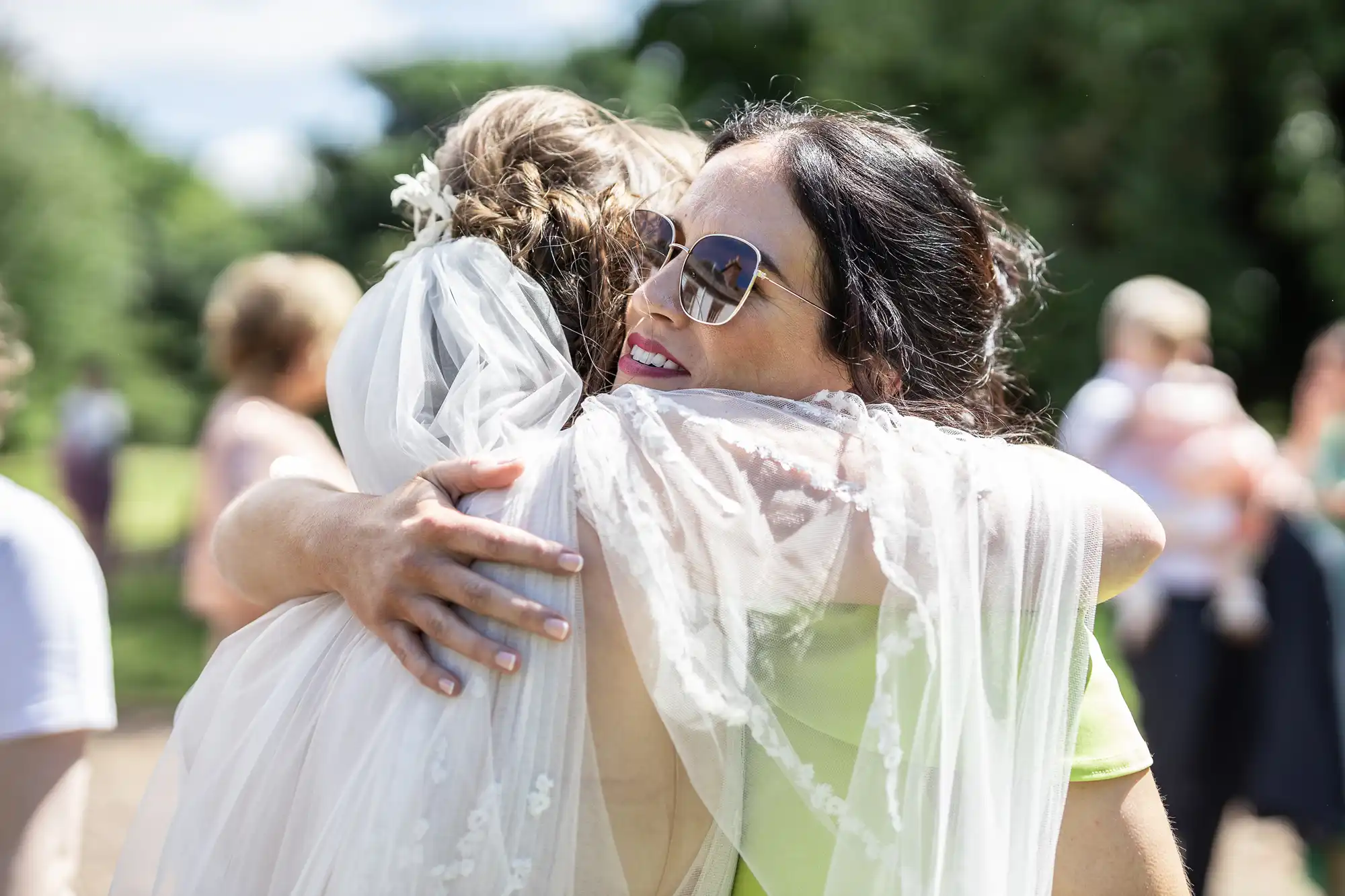 Two women embrace outdoors; one wears a white wedding dress and veil, the other wears sunglasses and a light green dress. The scene is sunny with blurred people and greenery in the background, conveying a joyful, celebratory moment.