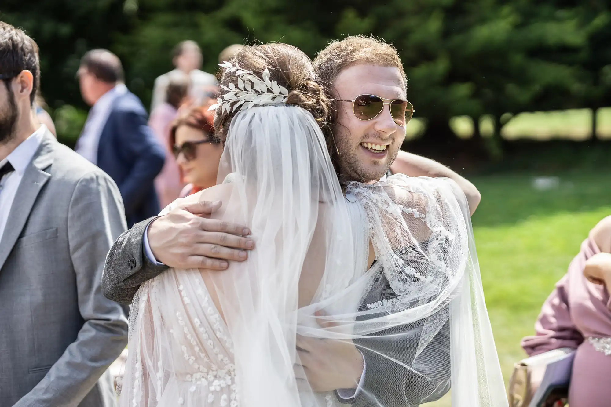 A smiling man in sunglasses hugs a bride wearing a white dress with a veil and floral headdress at an outdoor event. People dressed formally stand in the background, with sunlight and greenery visible around them.