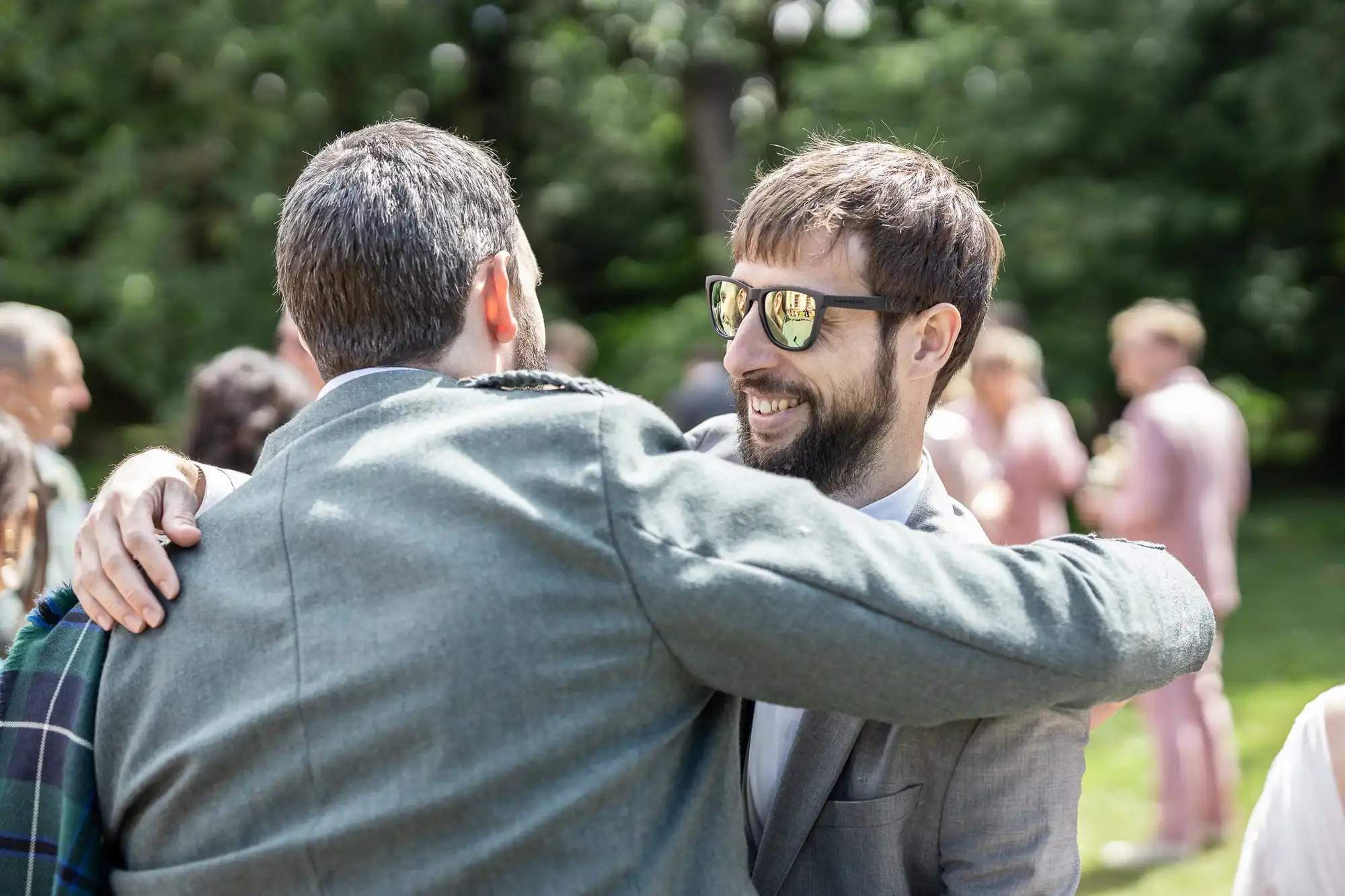 Two men embrace outdoors at a social event. One man, wearing sunglasses and a suit, smiles broadly. The other’s back is to the camera, wearing a grey jacket with a tartan detail. Other people and greenery are visible blurred in the sunny background.