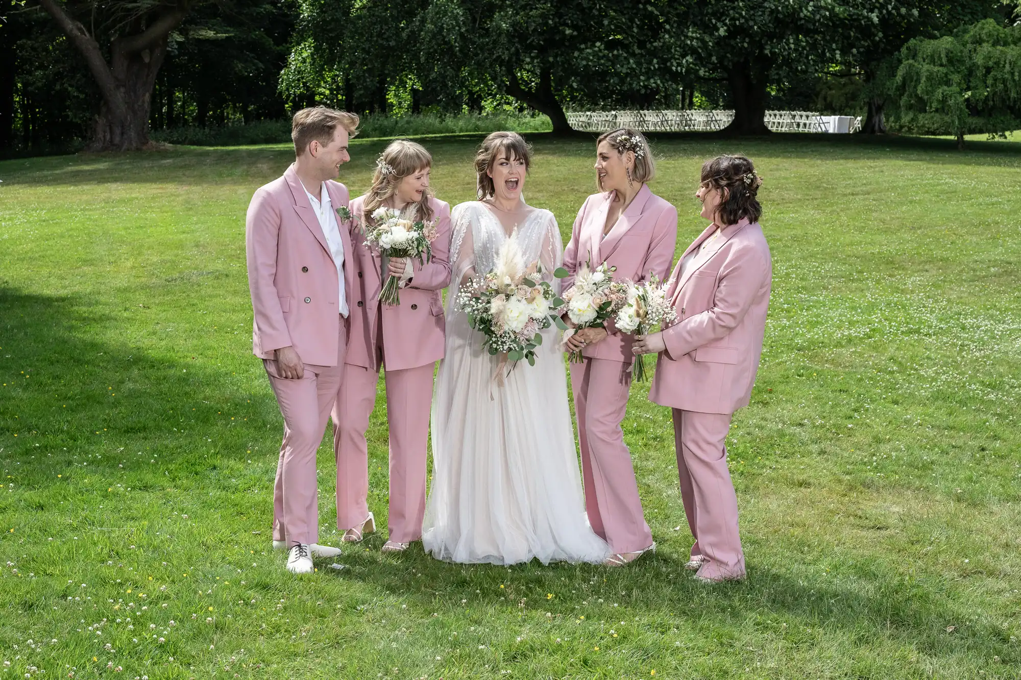A bride in a white gown stands smiling outdoors with four people in matching light pink suits. They hold bouquets of white flowers and greenery, standing on lush grass with trees in the background, appearing joyful and celebratory.