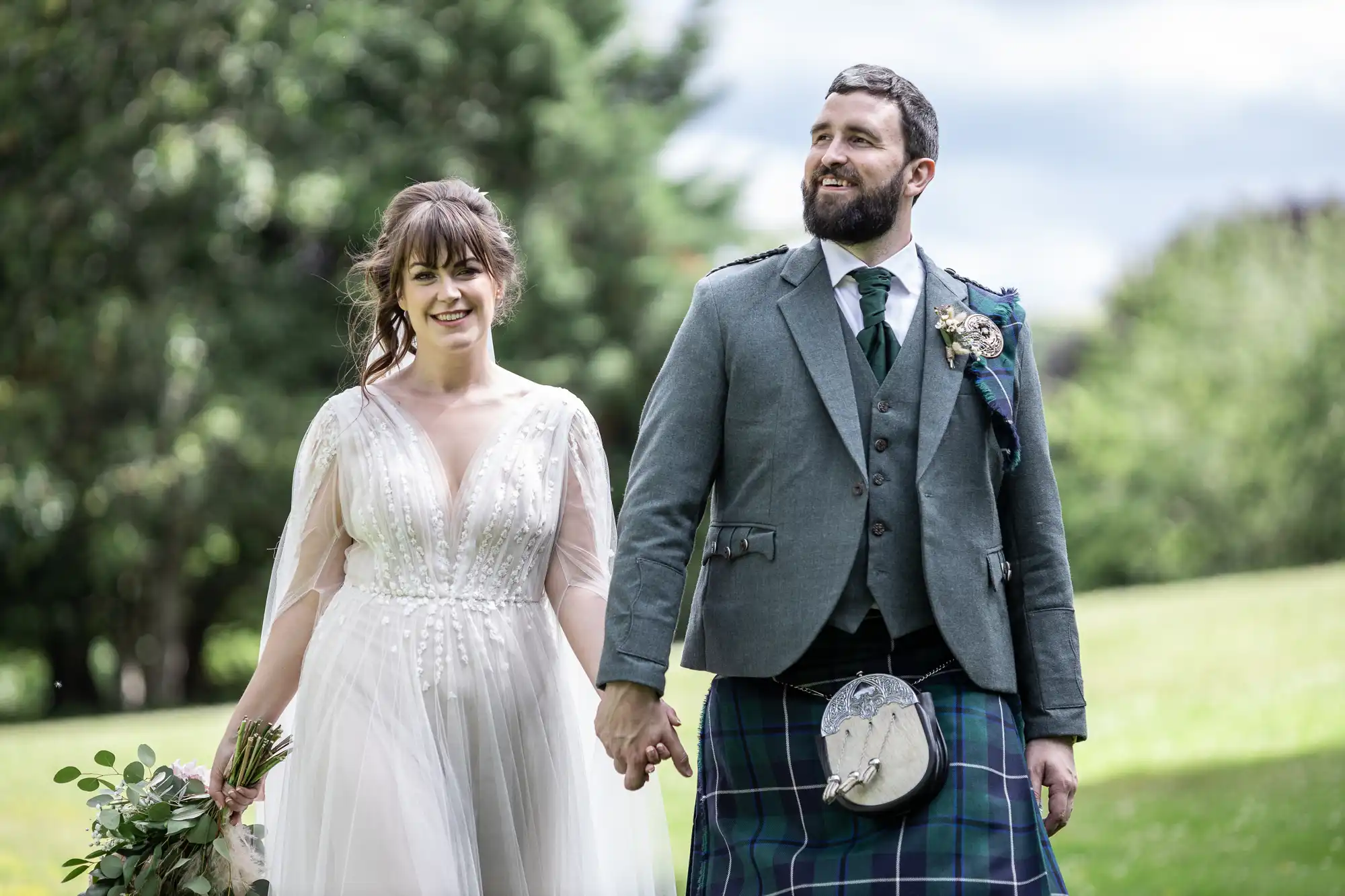 A bride in a flowing white dress with floral details holds a bouquet and smiles beside a groom in a grey jacket, checked kilt, and sporran. They hold hands and walk outdoors on lush green grass, with trees and a partly cloudy sky behind them.