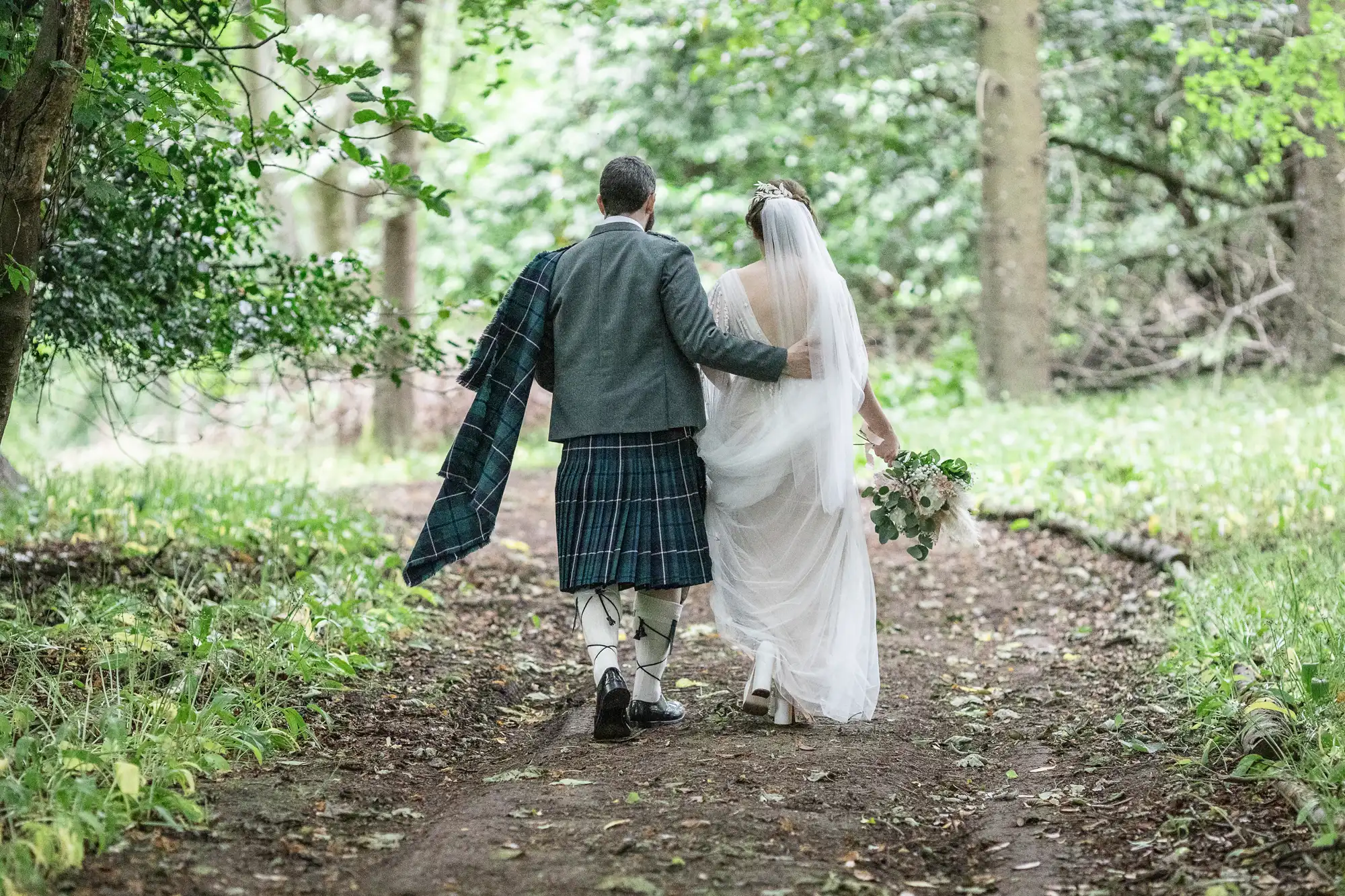 A bride in a white dress and veil walks arm-in-arm with a groom in a grey jacket and tartan kilt along a wooded path, surrounded by lush greenery. The bride holds a bouquet, and both are seen from behind, creating a peaceful, romantic scene.
