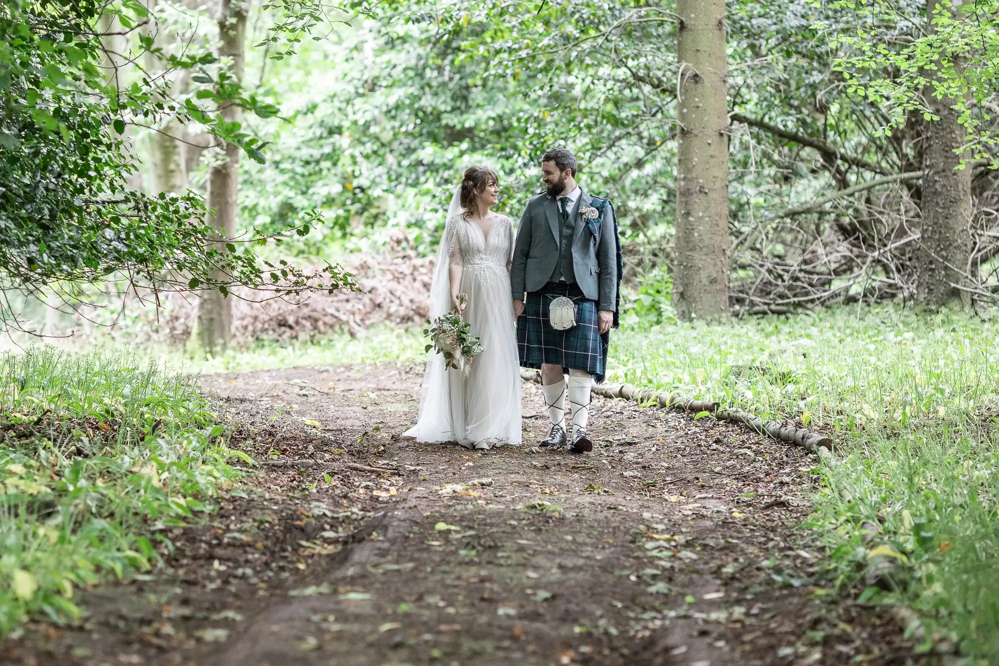 A bride in a long white dress holds a bouquet while walking hand-in-hand with a groom in a kilt and jacket. They stroll on a dirt path through a lush, green forest, looking at each other and smiling. Sunlight filters softly through the trees.