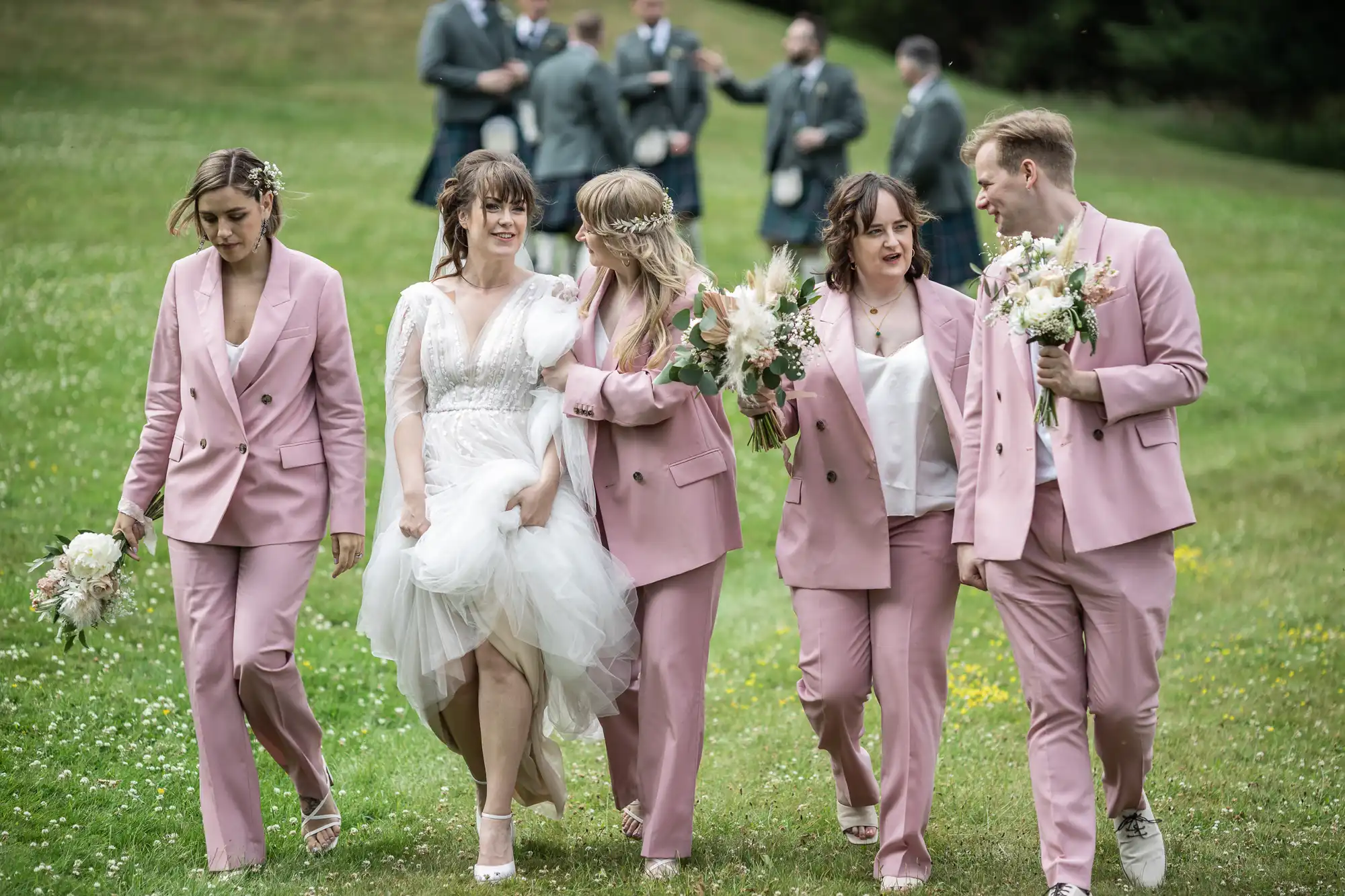 A bride in a white dress walks on grass, surrounded by bridesmaids and a groomsman in matching light pink suits holding bouquets. In the blurred background, men in grey kilts and jackets stand together. Everyone appears joyful and engaged in conversation.