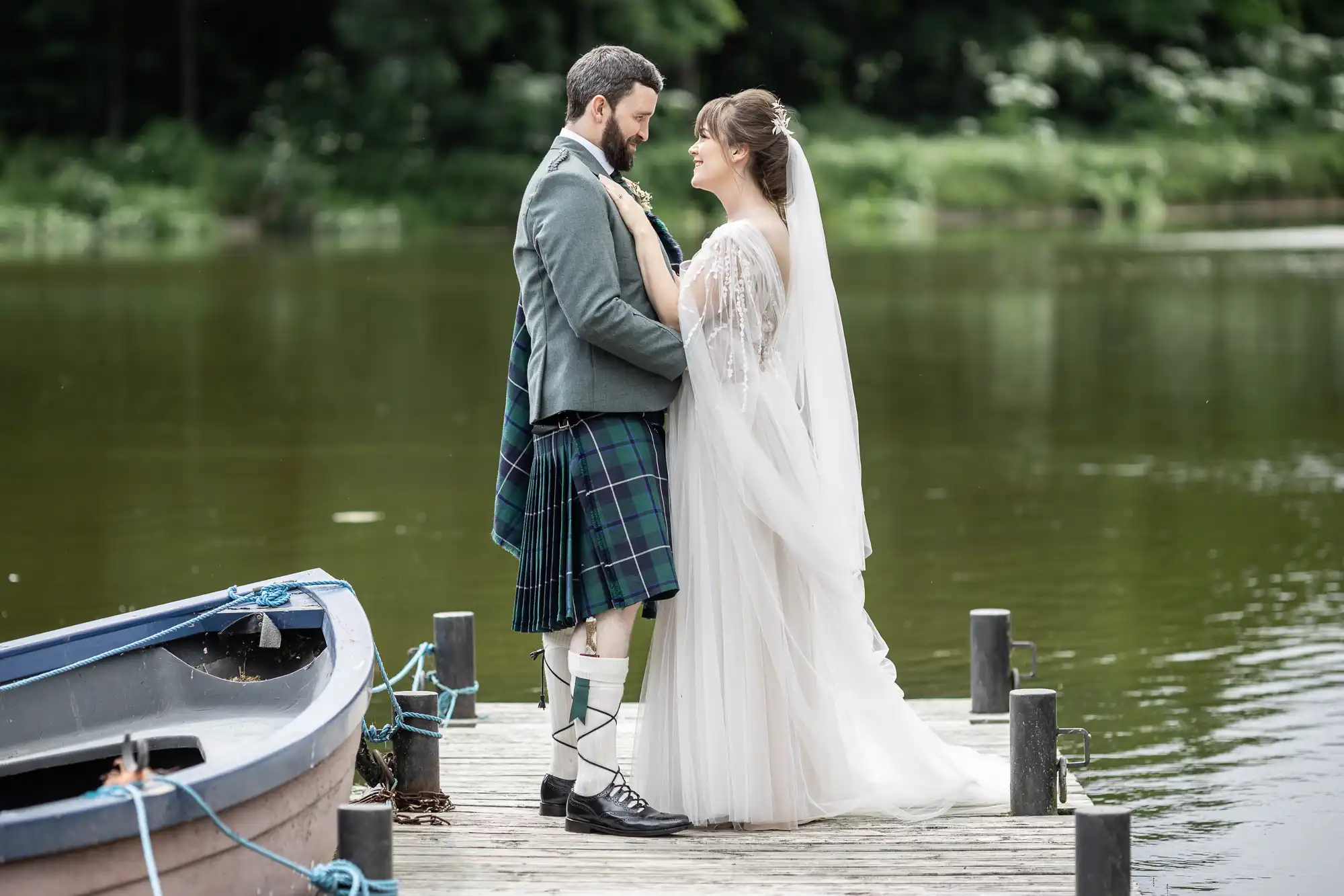 A bride in a white dress and veil stands on a wooden jetty by a lake, holding hands and gazing at a groom in a green jacket, tartan kilt, and knee-high socks. A small boat is tied to the jetty; green trees are visible across the water.