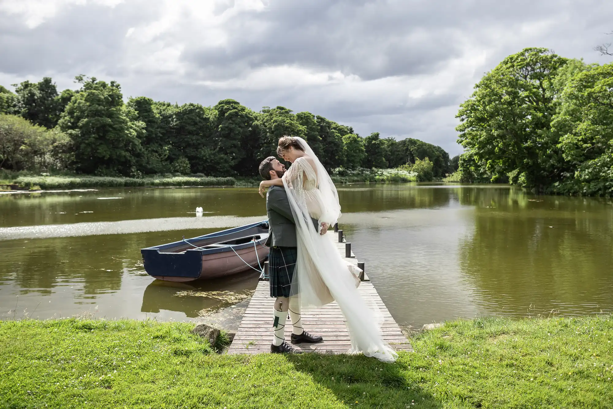 A groom in a kilt lifts a bride in a white dress and veil on a wooden jetty beside a calm lake, with green trees and a small blue rowing boat in the background under a cloudy sky.